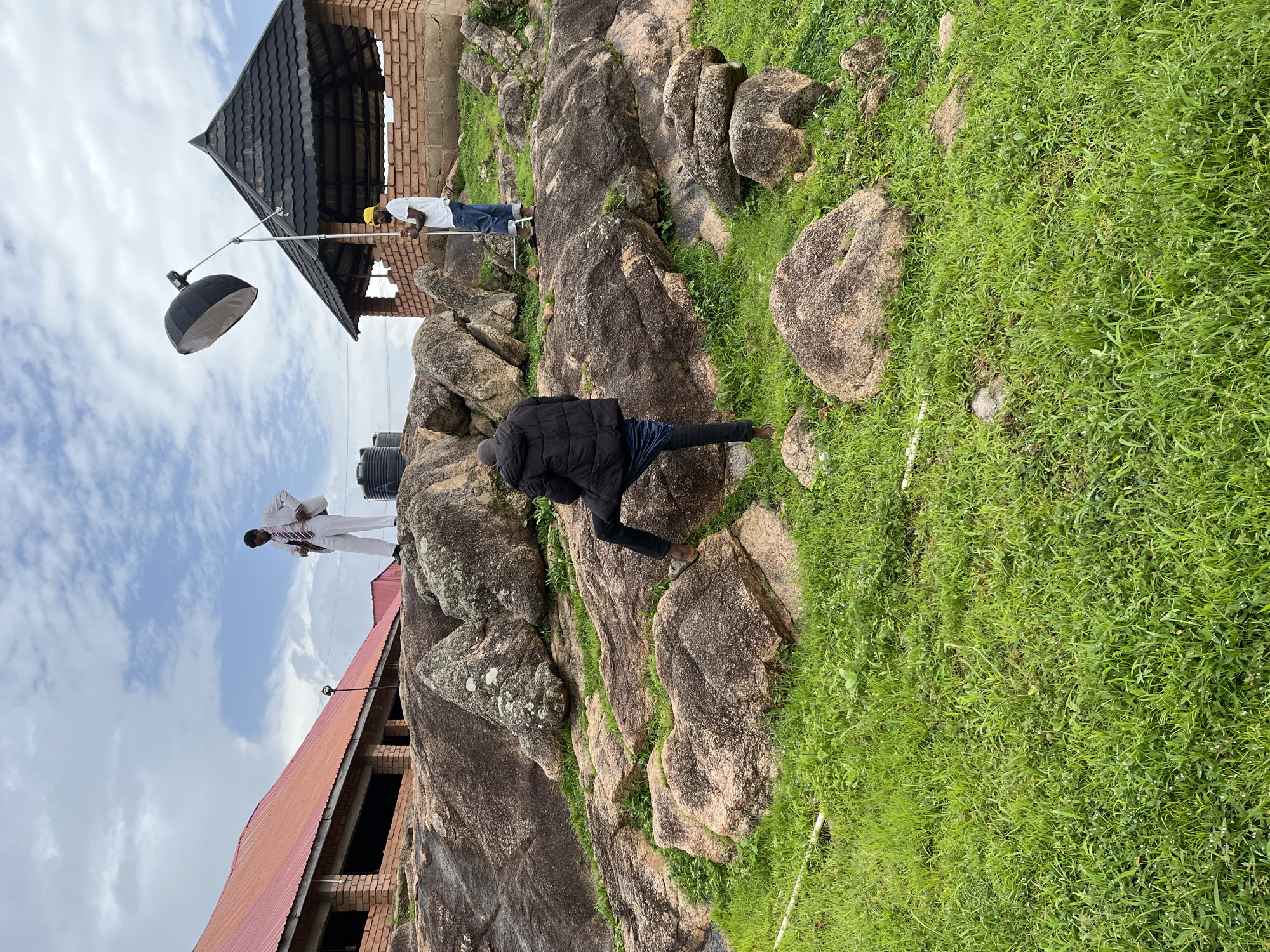 The media file depicts an outdoor scene with several subjects and objects amidst a rocky, grassy landscape. In the foreground, a person wearing a black jacket and dark pants is crouched on a large rock, appearing to be engaged in some activity. To the left, a person in a white shirt and jeans is standing next to a lighting setup, which includes a large parabolic reflector. Further to the left, another individual in a white suit with a striped tie is standing on a rock formation, looking towards the camera.
In the background, a partially constructed building with a red roof is visible. Adjacent to the building, a dark-colored water tank can be seen. The environment features large, weathered rocks interspersed with lush green grass, suggesting a natural, possibly hilly terrain. The sky is cloudy, with patches of blue, indicating daytime. The overall impression is that of an outdoor photoshoot or filming session taking place in a semi-developed area. The location cues, such as the brickwork and architectural style of the building, combined with the rocky landscape, are consistent with the provided context of Jos, Nigeria. No visible text is present in the image.