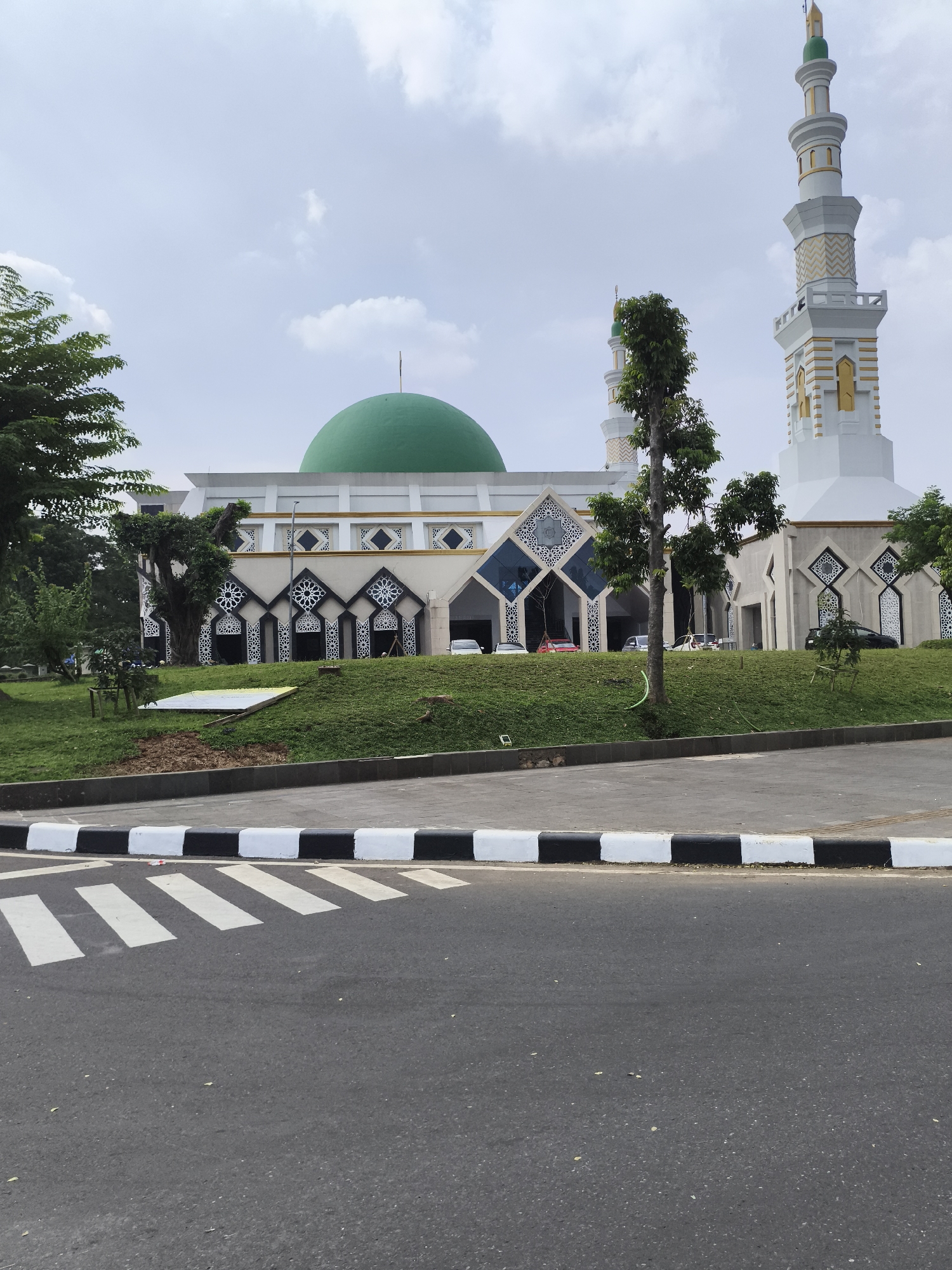 The image displays a large, modern mosque located in Cibinong, Indonesia, captured during daytime under a partly cloudy sky. The mosque is distinguished by its prominent green dome, centrally positioned on its roof. It features two tall minarets; the one on the right is notably taller and more elaborate, both sharing a design of white with intricate gold detailing and green-tipped finials.
The mosque's facade is predominantly white, decorated with striking geometric patterns in black and white, incorporating star and diamond motifs, accented with gold trim along its cornices. In front of the mosque, there are well-maintained, gently sloping grassy areas and several trees of various sizes, contributing to a lush, green landscape. A few cars are partially visible, parked near the mosque's lower entrances.
The foreground of the image shows an asphalt road. A black and white painted curb separates the road from the mosque grounds, and a clearly marked zebra crossing is visible on the asphalt, indicating a pedestrian path. No people are present in the scene, and there is no visible text. The overall atmosphere appears peaceful and serene.