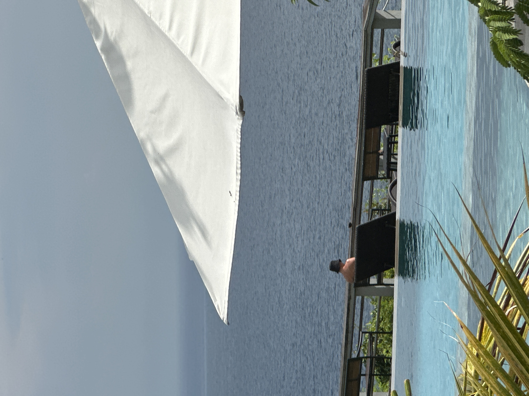 This image, taken in Kuta Selatan, Indonesia, depicts a tranquil outdoor resort scene under bright daylight. The composition is split diagonally by a large white umbrella, partially visible in the upper left, providing shade.
In the middle ground, a vast expanse of calm, deep blue ocean stretches towards the horizon. In front of the ocean, on an elevated patio or balcony, a lone individual is seated in a dark outdoor chair, facing away from the viewer and towards the sea. The person wears a dark hat, suggesting a moment of relaxation and contemplation of the view. Other dark outdoor furniture, including additional chairs and tables, is partially visible on this lounging area.
Dominating the lower right portion of the image is an infinity pool with clear, light blue water, its edge seemingly merging with the ocean beyond. The surface of the pool subtly reflects the surroundings. In the extreme foreground on the lower right, vibrant green and yellow-green foliage from a tropical plant adds a natural element to the scene. The sky above the ocean is a clear, light blue, indicative of fair weather. The overall ambiance is one of peace and leisure in a scenic tropical location. No text is visible in the image.
