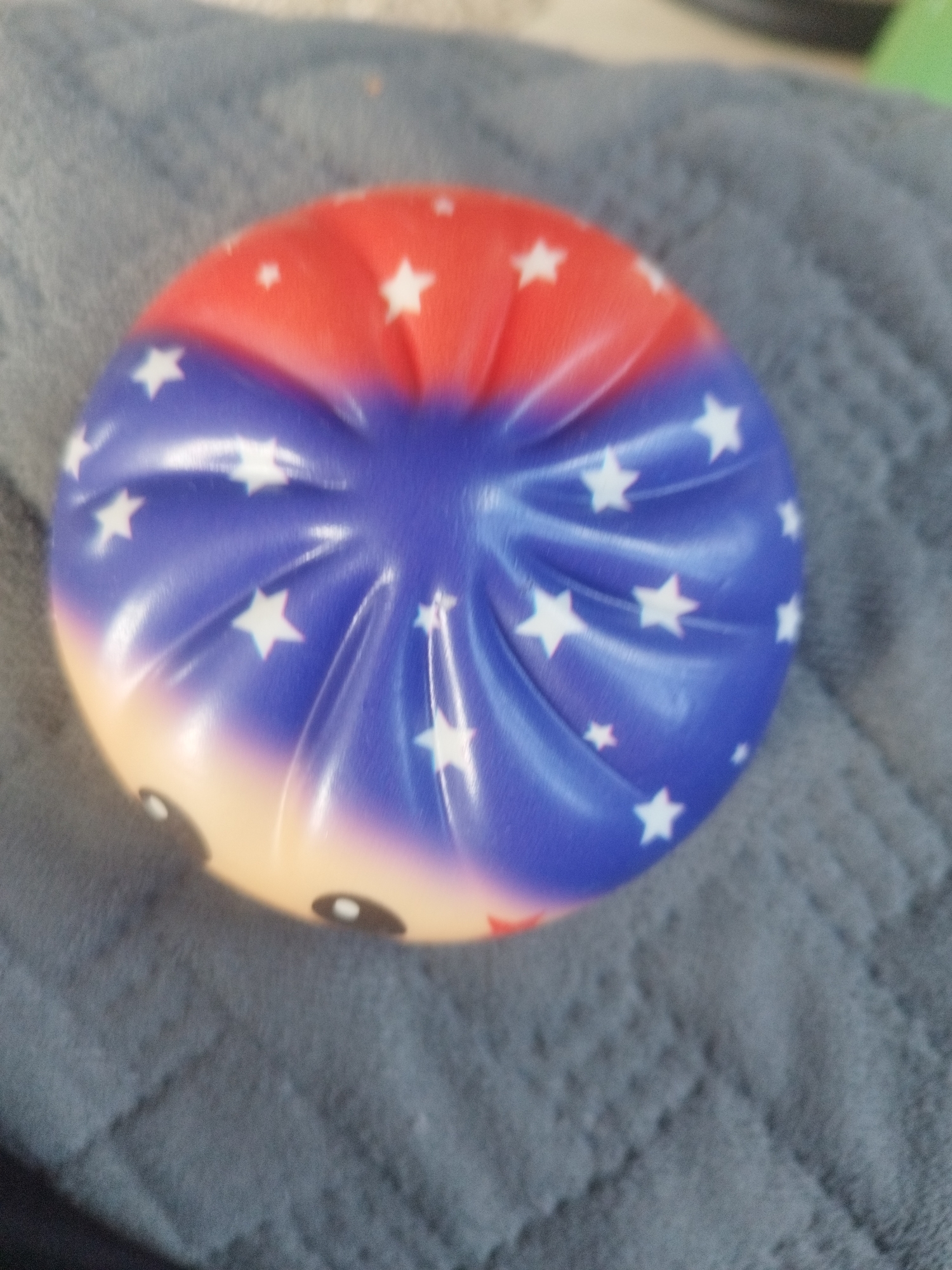 A close-up, slightly angled shot captures a patriotic-themed, circular, squishy toy resting on a dark gray, textured fabric. The toy is divided into three color sections: red at the top, fading into blue, and then a pale yellow at the bottom. White stars are scattered across the red and blue sections. The blue section has a rippled texture, creating shadow and highlight effects. The pale yellow section appears to depict a face, with two small, dark, circular eyes with white pupils visible. The fabric beneath the toy appears to be a quilted blanket or similar material with a soft texture. The background is out of focus and shows a dark, indistinct area. There is no indication of the location within the city of Oskemen, Kazakhstan.
