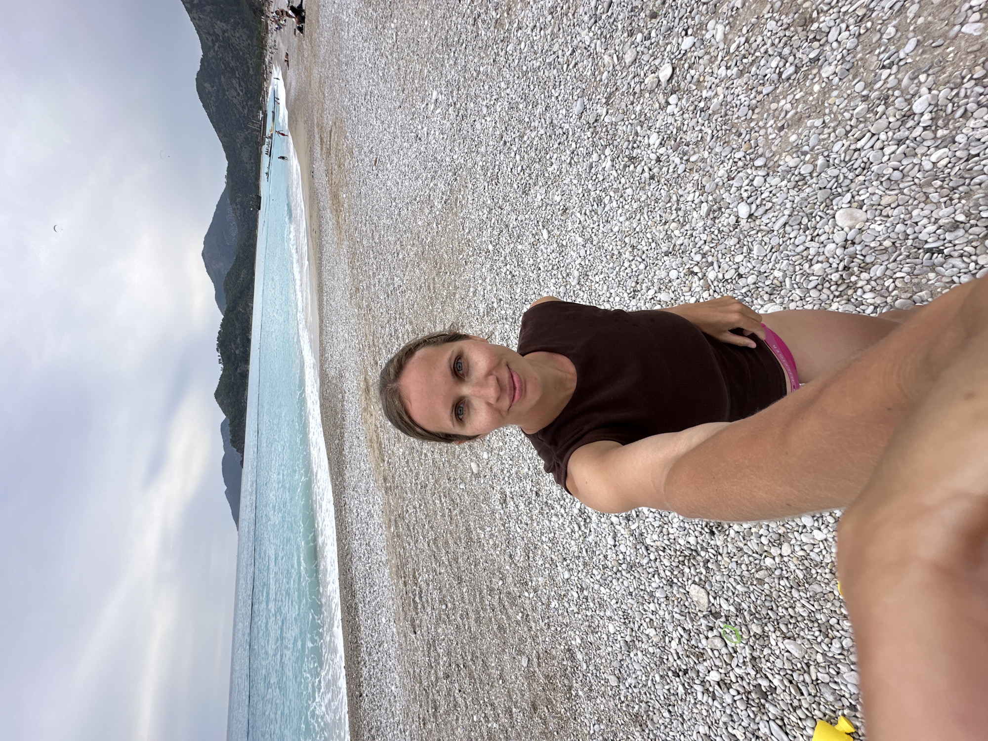 The image is an eye-level, outdoor selfie of a woman on a pebble beach in Fethiye, Turkey. The woman is fair-skinned with blonde hair and is wearing a dark brown t-shirt and pink shorts. She is lying on the beach with her arm extended to take the photo, and a portion of her bare legs are visible. The beach is made of small, white and gray pebbles. In the background is a turquoise sea with small waves and a mountain range. The sky is overcast. There are people on the beach in the distance. A green rubber band and yellow cloth are visible near the woman's leg on the stones.
