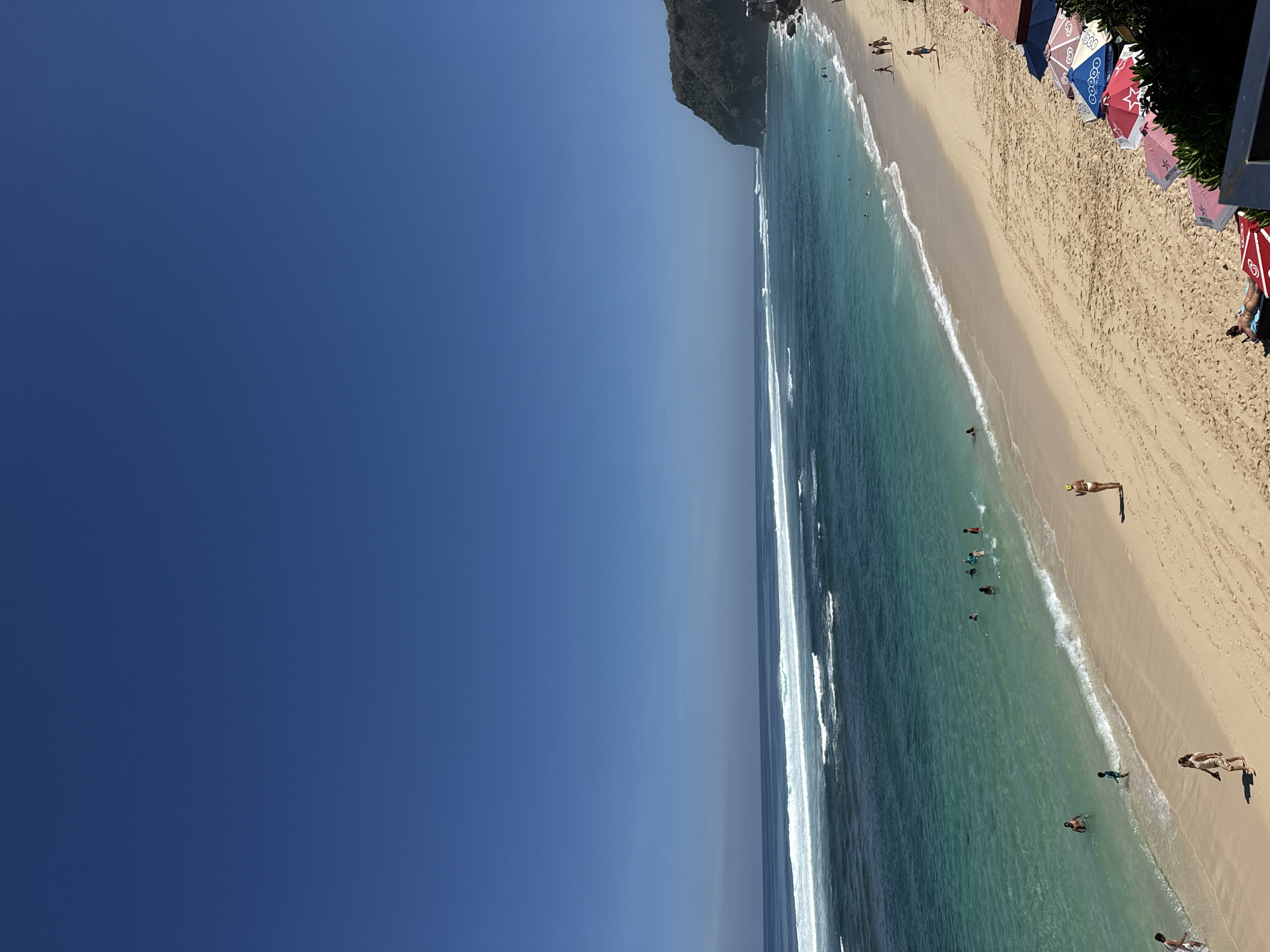 The image depicts a beach scene in Kuta Selatan, Indonesia, under a clear blue sky. The ocean exhibits shades of turquoise and deep blue, with white waves breaking near the shore. A sandy beach stretches along the coastline, populated by several individuals. Some people are in the shallow water, swimming or wading, while others are walking on the beach. A prominent cliff face covered in vegetation rises on the right side of the frame, bordering the beach and the sea. A row of colorful beach umbrellas is set up on the sand near a building structure. The overall atmosphere is one of a sunny day at a tropical beach.