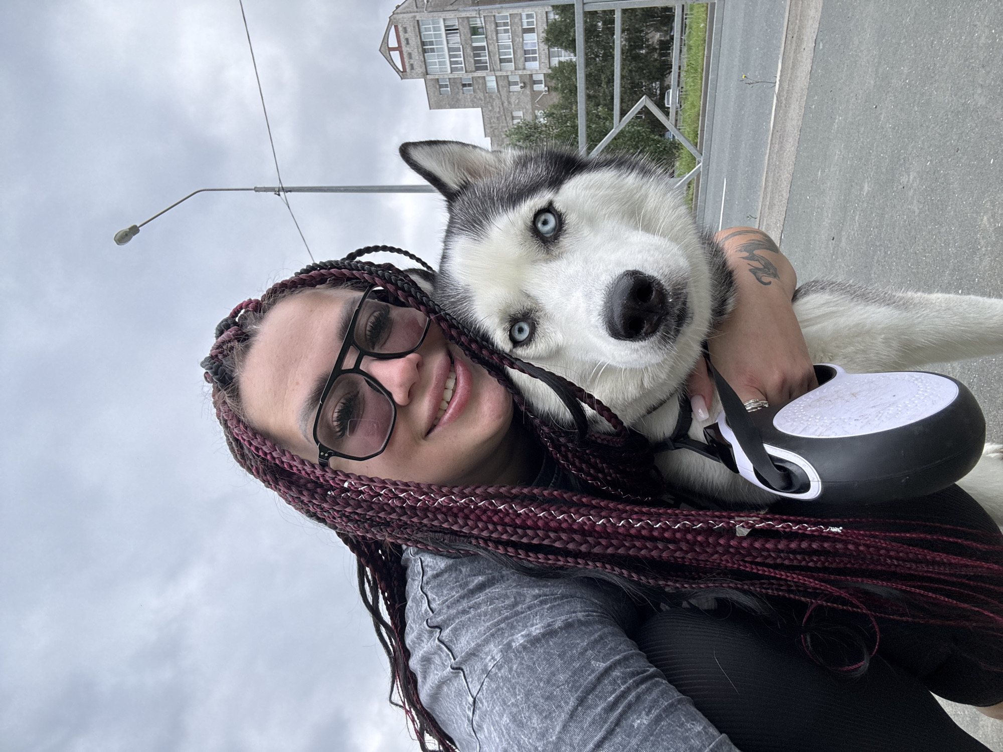 A medium shot shows a woman with dark red braids and glasses posing for a selfie with a husky dog. The woman is wearing a gray t-shirt and black leggings. She is holding the dog by its leash, which is a retractable black and white model. The dog is a black and white husky with striking blue eyes. They are outdoors in a city setting. In the background, there is a tall building with balconies, trees, a street lamp, and a grey sky. The ground is paved. The image appears to be taken in the city of Tolyatti, Russia, possibly on a street or sidewalk, as indicated by the urban elements and architecture.