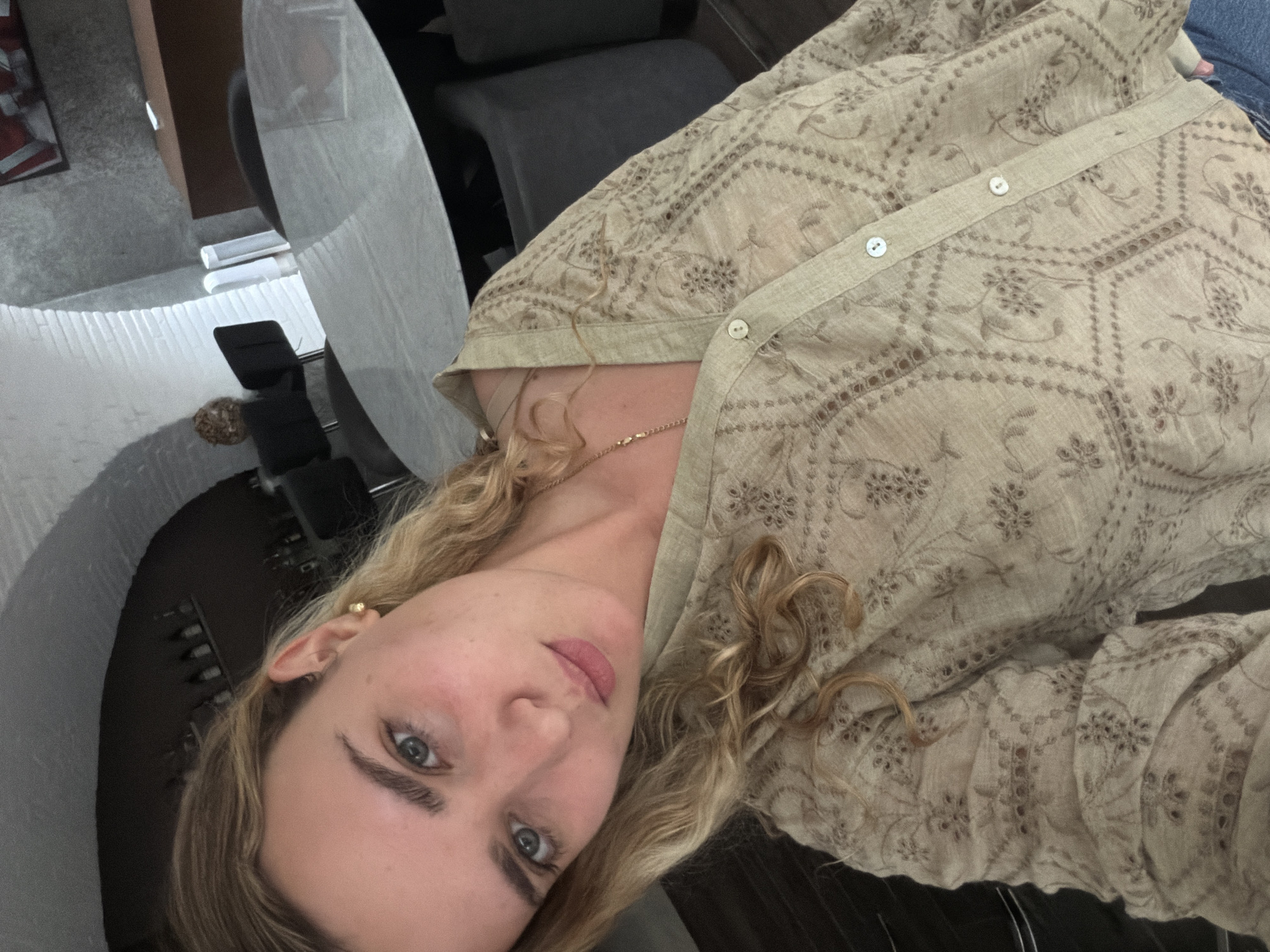 A woman with blonde curly hair, wearing a patterned beige button-down shirt, is the primary subject of the image. She is looking directly at the camera with blue eyes. A gold chain necklace is visible around her neck. The background includes a white round table, a grey chair, and what appears to be a room with various objects. The location cannot be confirmed as San Bernardino Tlaxcalancingo, Mexico, based solely on the visual information provided.