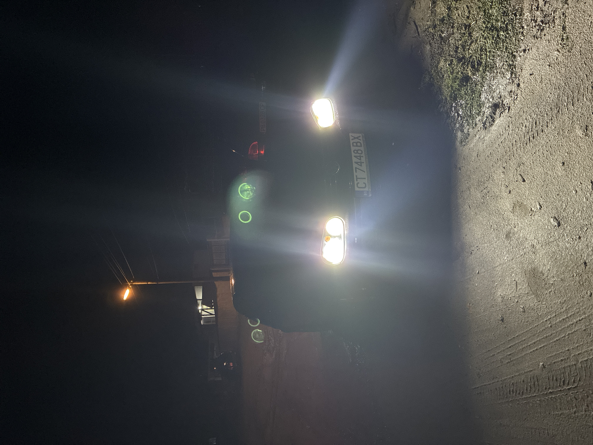 The image depicts a dark outdoor scene, likely at night, in an unknown city. A car with illuminated headlights and tail lights is parked on a paved surface, likely a road. The car's license plate reads "CT 7448 BX." The road surface has tire tracks visible. Adjacent to the road, there is a patch of vegetation. In the background, a street light is visible. There is a building or structure behind the car. Another license plate, "PB 0326 CP" can be seen behind the car.