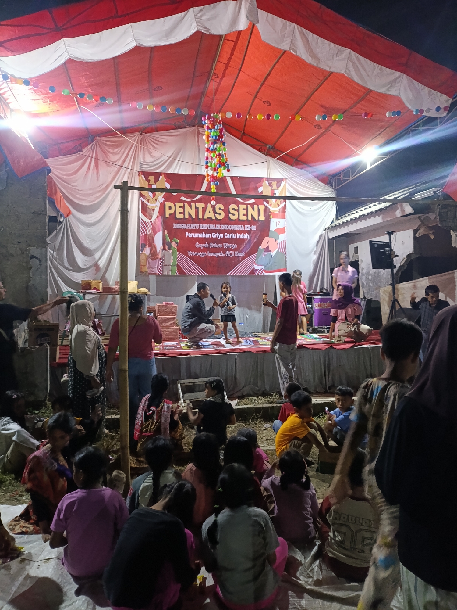 The image depicts an outdoor evening event in Cariu, Indonesia, likely a community gathering or cultural performance. A temporary stage is set up under a large red and white tent, adorned with colorful string lights and a beaded chandelier. A banner behind the stage reads "PENTAS SENI" (Art Performance) and "DIRGAHAYU REPUBLIK INDONESIA KE-71" (71st Anniversary of the Republic of Indonesia), indicating the event is celebrating Indonesia's independence.
On the stage, a man is kneeling and speaking into a microphone, facing a young girl who is standing and also holding a microphone. Behind them, several people are present, and boxes are stacked on shelves. In the foreground, a crowd of people, predominantly children, are seated on the ground, facing the stage. They appear to be watching the performance. Some adults are also visible, standing and observing the event. The environment suggests a casual, communal atmosphere, with the ground covered in tarps or mats for seating. The lighting is dim, with stage lights and string lights illuminating the immediate performance area, while the surrounding area is darker.