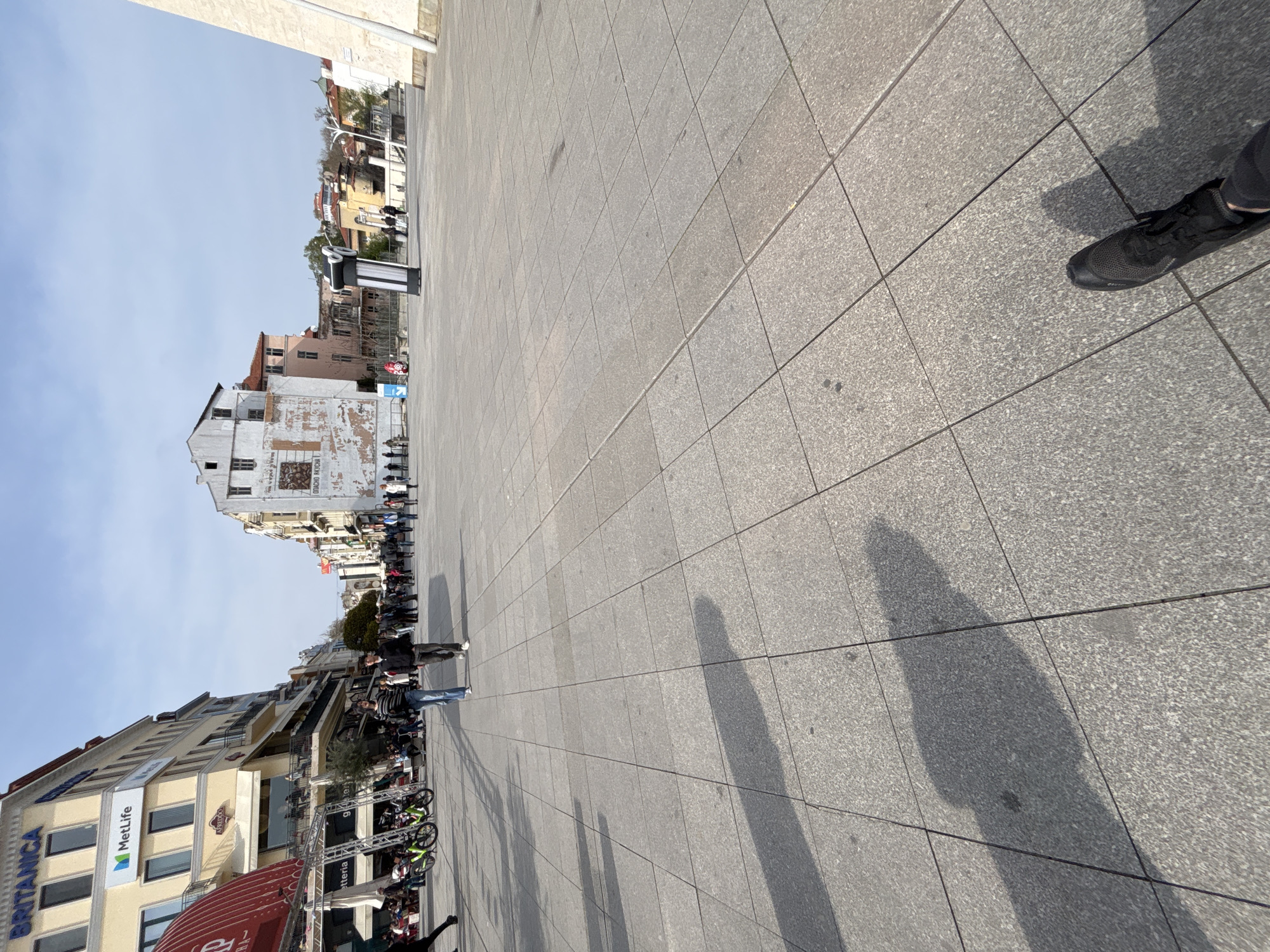 The image depicts a pedestrianized square in Plovdiv, Bulgaria. The square is paved with grey stone tiles. On one side is a line of buildings with shops and restaurants, including a building with "BRITANICA" and "MetLife" signs. On the opposite side is a row of older buildings, some with visible signs of wear. A line of people is visible, likely waiting for something. There are bicycles and event-related structures nearby, possibly indicating a sporting event or gathering. The sky is clear and blue. A person's shoe can be seen in the corner of the photo, along with a shadow across the square.