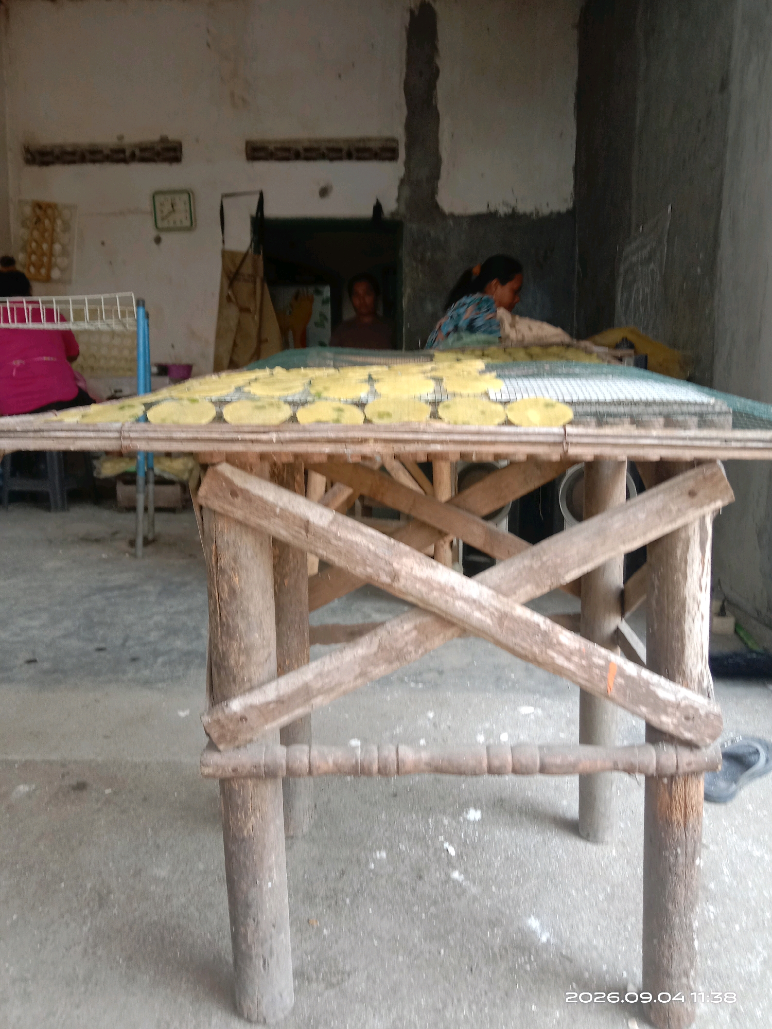 The image depicts an outdoor scene in what appears to be a rural or semi-urban area of Sepanyul, Indonesia. In the foreground, a rustic wooden structure resembling a table or drying rack is prominent. This structure is made of rough-hewn logs and planks, with cross bracing for stability. Resting on top of this structure is a mesh rack covered with numerous circular, flat objects that appear to be drying, possibly food items like crackers or chips. The color of these items is a pale yellow.
In the background, a building with unfinished concrete walls is visible. To the left, a person wearing a bright pink garment is partially visible, seated near a white rack that holds multiple items, potentially eggs. Further back, another person, a woman with dark hair tied back, is visible in profile, working with the drying items. She is wearing a patterned blue and green top. A clock is mounted on the wall of the building to the left. The ground appears to be a concrete or dirt surface. The overall impression is of a food production or drying process taking place in an open-air environment.