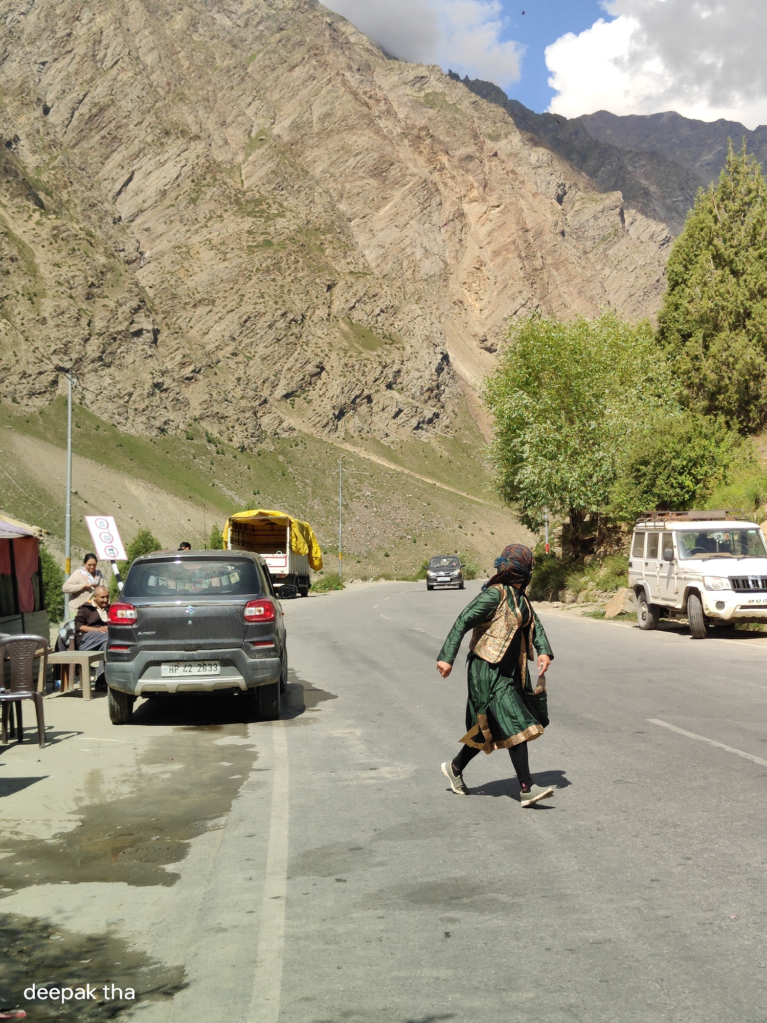 This is a medium-shot, eye-level, outdoor photograph taken in Darcha, India. The image depicts a woman in traditional clothing, a green dress with gold trim, and a patterned headscarf and vest, walking across a paved road. She is in motion, with her legs bent as if mid-stride, and a bag is slung over her right shoulder. To her left, a dark grey car with a license plate reading "HP 42 2633" is parked. Further to the left, a yellow-covered truck is stopped. Behind these vehicles, a person is seated on a bench next to a small roadside structure. A signpost with circular speed limit indicators (40, 50) is visible near the parked car. In the background, a steep, rocky mountain with sparse green vegetation dominates the landscape. Several trees are scattered on the mountainside and along the road. A white SUV is parked on the right side of the road, facing away from the viewer. The sky is partly cloudy with blue patches. The road appears wet in places, suggesting recent rain or a water source. The overall scene conveys a sense of travel and rural mountain scenery.