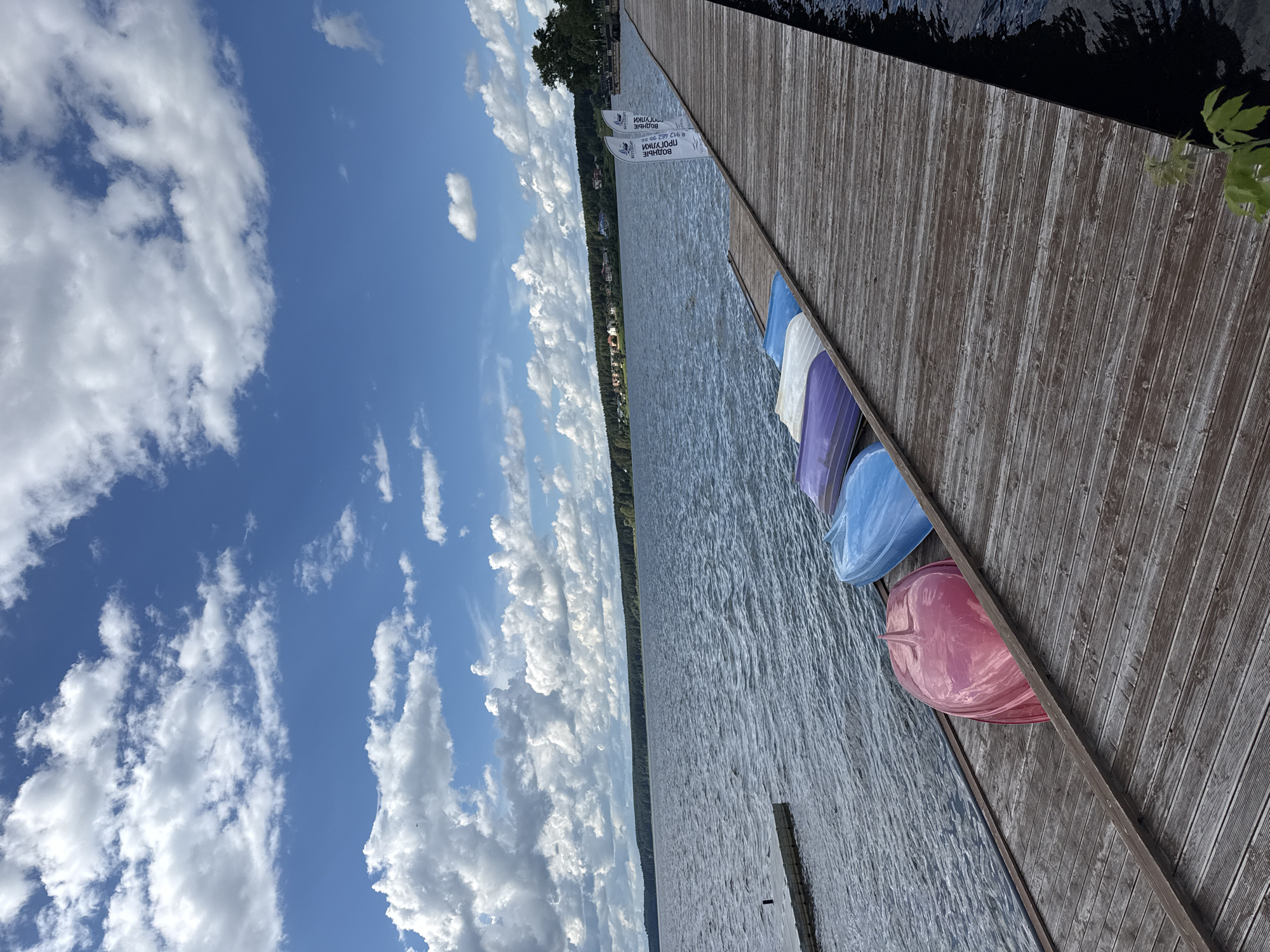 This image captures a serene waterside scene with a wooden pier extending into a calm body of water. Several colorful boats, including a bright pink one, are neatly lined up on the pier, suggesting a recreational area. The background reveals a lush, tree-lined shore with a scattering of houses, indicative of a settlement along the water's edge. The sky is a vibrant blue, adorned with a dramatic display of fluffy white clouds, hinting at fair weather. The scene appears to be located in the city of Votkinsk, Russia, a place known for its picturesque natural landscapes and connection to the water. There are no people visible in the frame, and the overall impression is one of peaceful tranquility and natural beauty.