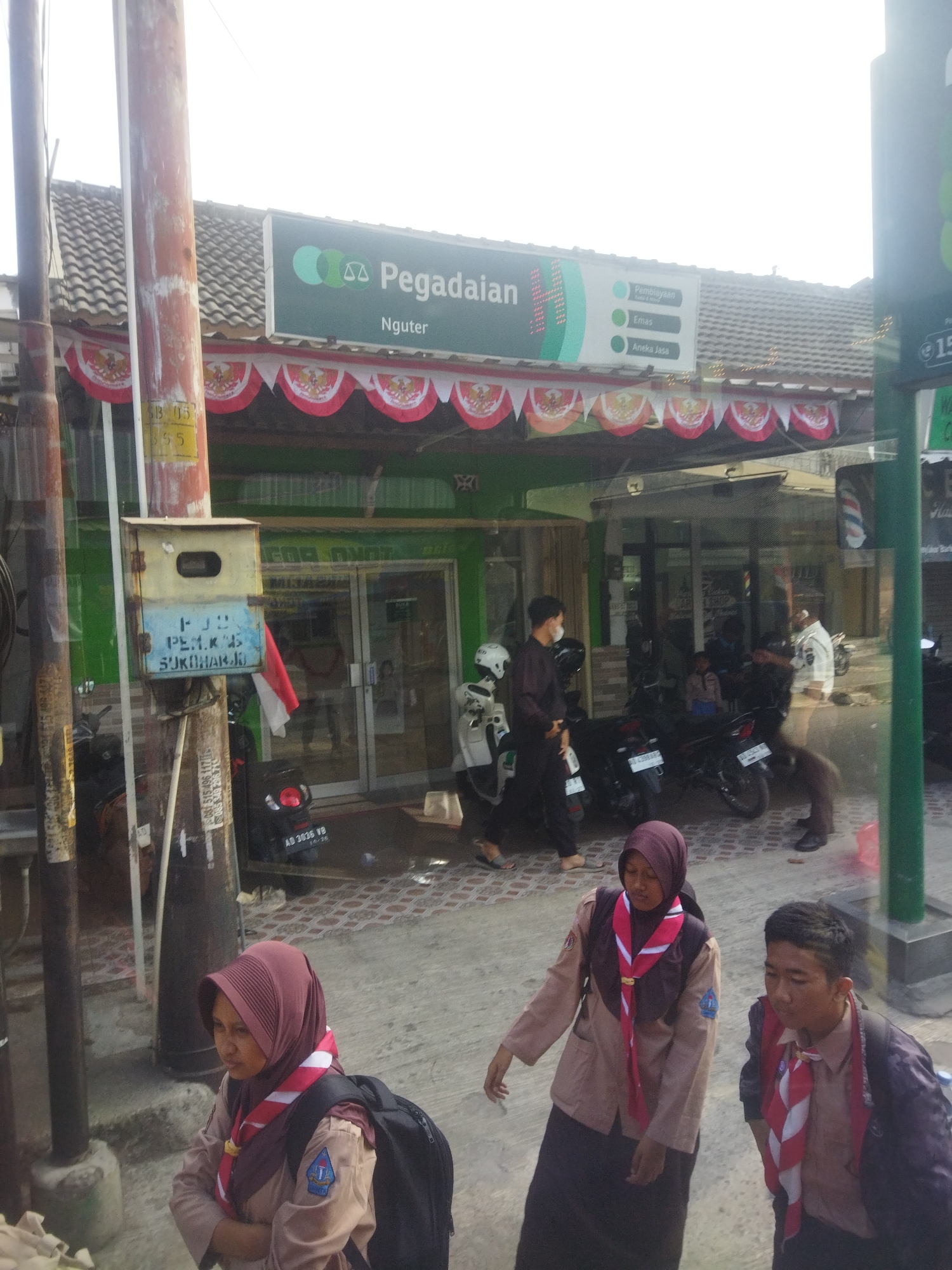 The image shows the exterior of a Pegadaian (pawnshop) branch in Nguter, Indonesia. A green awning with the Pegadaian logo and text "Pegadaian Nguter" is visible above the entrance. Below the awning, a string of red and white Indonesian flags is displayed. The entrance to the building appears to be glass doors. In front of the building, several motorcycles are parked. Three young people in Scout uniforms are walking on the sidewalk in the foreground. One person is looking to the left, while the other two are looking forward. A utility pole with a blue and yellow box attached is on the left side of the image. The ground is paved with concrete and tiles. The overall scene suggests a busy street in a commercial area.