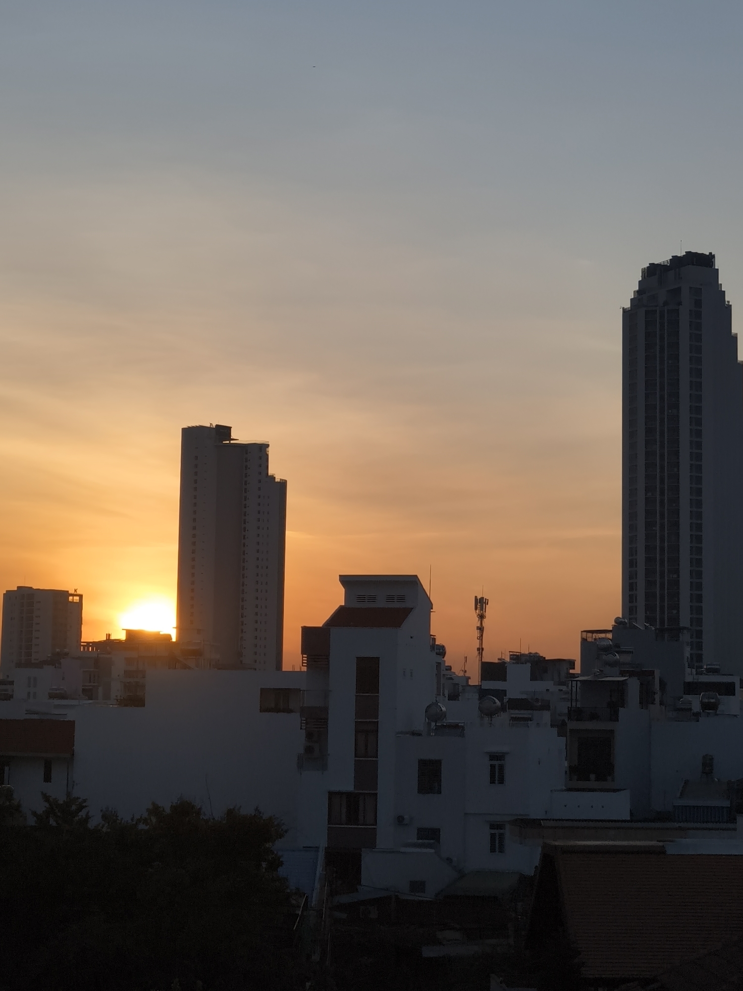 This is a vertical, medium-shot photograph of a city skyline at sunset. The sun, a bright orb, is partially obscured by buildings on the left side of the frame, casting a warm, golden-orange glow across the sky. The sky transitions from this vibrant orange near the horizon to a pale blue at the top of the image. Silhouetted against the sunset are several modern buildings of varying heights.
Two tall, slender skyscrapers are prominent. One is in the center-left, and another, slightly taller and more detailed with a stepped roofline, is on the far right. A shorter building with a distinctly layered top is located in the mid-ground, slightly to the right of the center. In the foreground and mid-ground, there are several lower-rise buildings, mostly white, with visible windows and rooftop structures, some of which appear to be air conditioning units or water tanks. A telecommunications tower with multiple antennas is visible between some of the buildings in the mid-ground. The lower portion of the image shows dark masses of trees, suggesting a park or residential area with foliage in the foreground.
The overall mood is serene and peaceful, capturing the beauty of a sunset over an urban environment. The strong contrast between the bright sky and the silhouetted buildings creates a dramatic visual effect. There is no visible text in the image. The time of day is clearly sunset, and the weather appears clear. The presence of skyscrapers and the dense arrangement of buildings suggest an urban setting, likely a city.