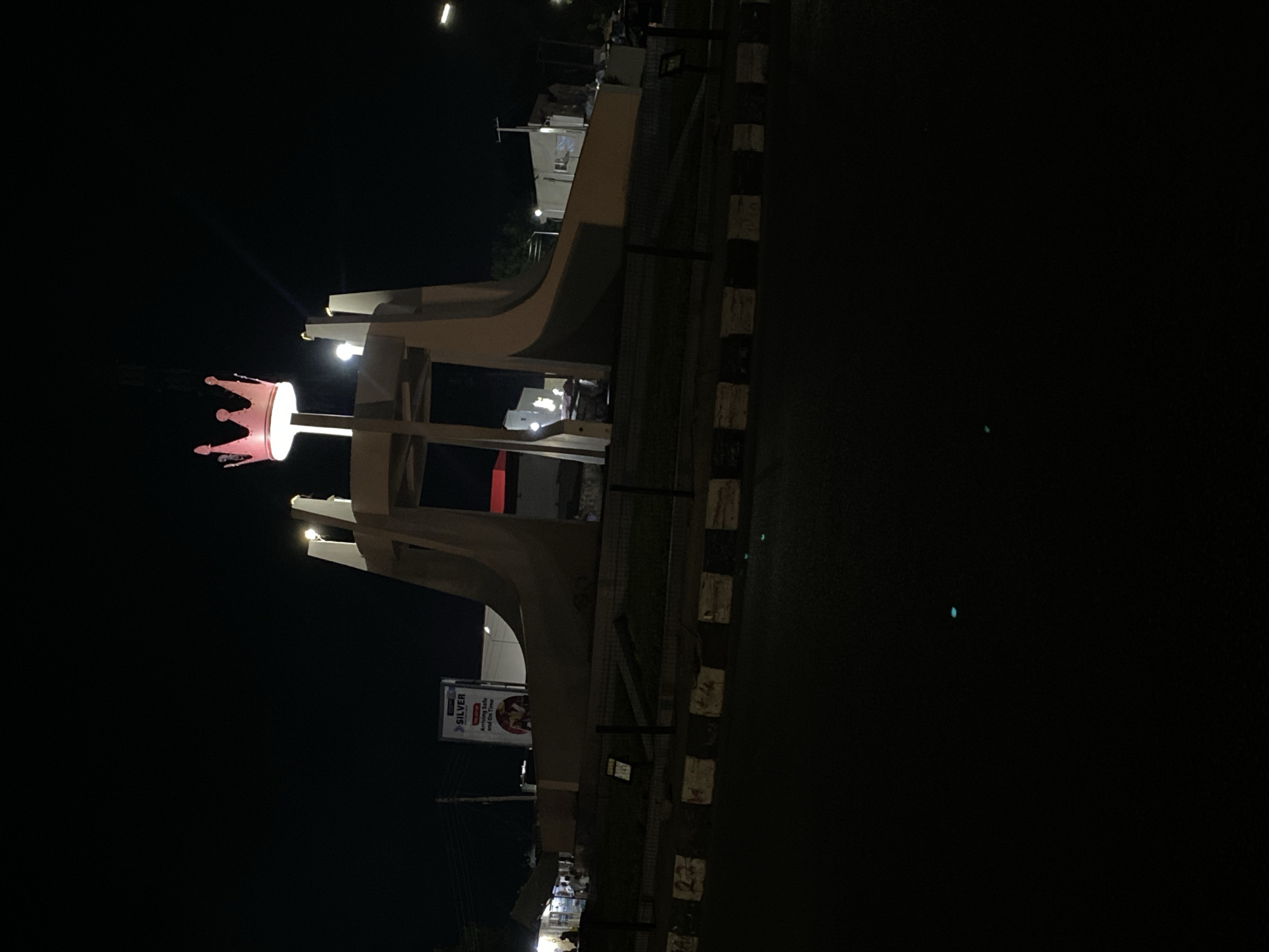 The image shows the illuminated coronation monument at night in Jos, Nigeria. The monument features a large pink crown on top of a white, stylized archway. A lit Silver advertisement is visible. The surrounding area is dark. There are no people clearly visible in the image.