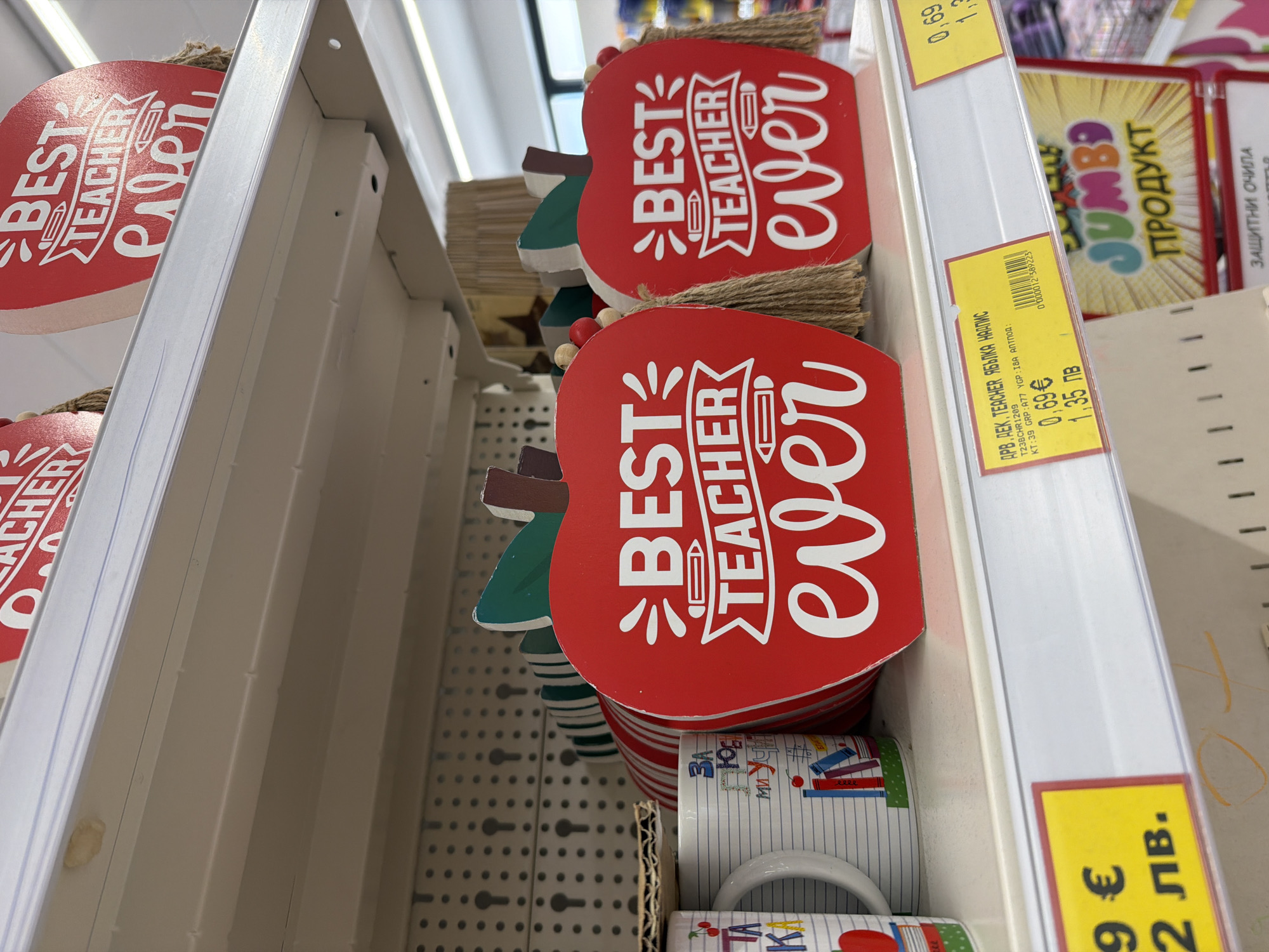 This is a medium shot, eye-level, indoor photograph taken at a retail store in Plovdiv, Bulgaria. The scene is a shelf display showcasing decorative apple-shaped items with the text "BEST TEACHER ever" printed in white on a red background. Two such apple decorations are prominently featured, one slightly to the left and the other to the right. Both have a stem and leaves at the top and are adorned with brown twine tassels and beads.
To the left of the apple decorations, a white mug with a striped pattern and illustrations of books and cherries is visible. Behind the mug, and partially obscured by the apple decorations, are other items, including what appears to be a rolled-up item with red and white stripes and a piece of corrugated cardboard.
Below the shelf with the apple decorations, a yellow price tag is visible on the left side of the image, displaying "9 €" and "2 лв.". To the right of this, another yellow price tag is present, showing "0,69 €" and "1,35 лв.". This price tag also contains a barcode and some text in what appears to be Bulgarian.
Further to the right and lower down, a display box with the word "JUMBO" in colorful letters and the text "ПРОДУКТ" (Product) below it is visible. The overall environment suggests a retail setting, likely a gift shop or a department store aisle. The lighting is bright, indicating artificial store lighting. No people are present in the frame.