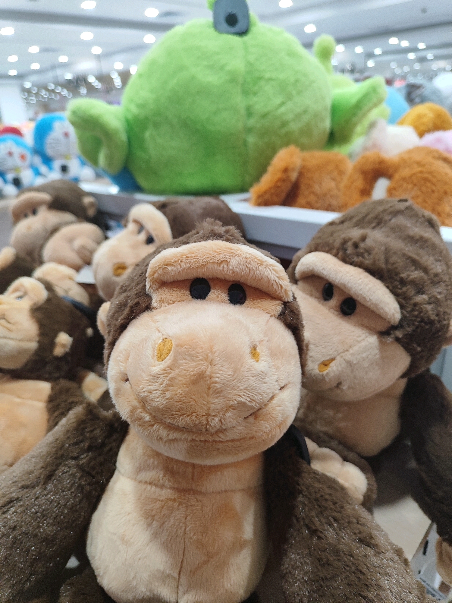In Sukowiryo, Indonesia, a close-up shot showcases a collection of plush stuffed animals, primarily focusing on several monkey figures. The monkeys are positioned at varying depths, with the one in the foreground taking center stage. This monkey, with its light brown and dark brown fur, has a smiling expression and black eyes. Its facial features include a lighter colored muzzle with yellow accents on its cheeks and nose.
Behind the prominent monkey, other similar monkey plush toys are visible, some partially obscured. In the background, a large, bright green alien-like plush toy is dominant. The setting appears to be a retail environment, possibly a toy store, with shelves and other colorful stuffed toys, including what appear to be characters from Doraemon, discernible in the blurred background. The lighting is bright, suggesting an indoor commercial space.