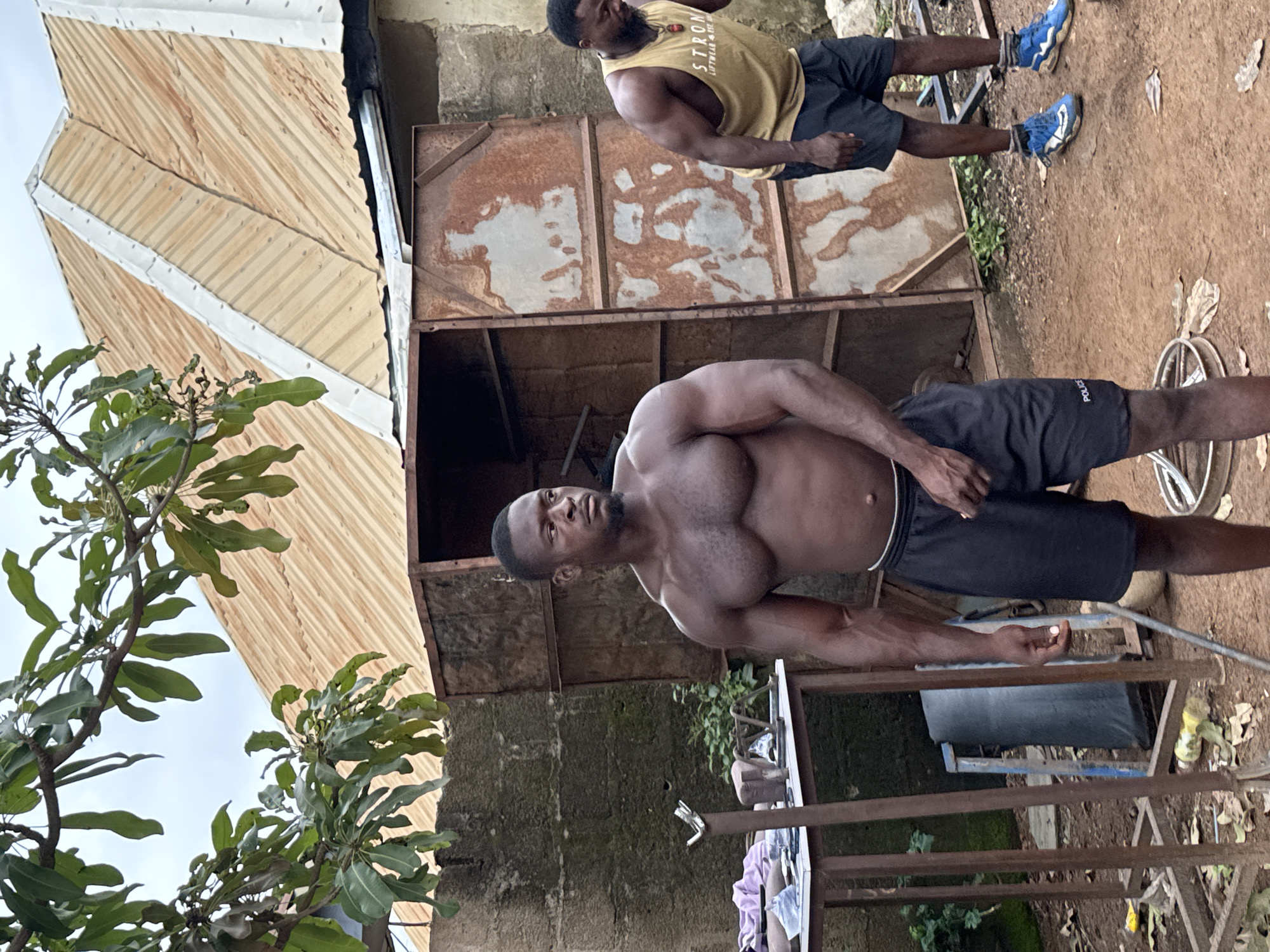 Two muscular Black men are standing outdoors in what appears to be a makeshift gym area. The man on the left is shirtless, wearing black shorts with "POLICE" written on the left leg, and is looking to the left. The man on the right is wearing a yellow tank top with text that appears to say "STRONG," black shorts, and blue athletic shoes. He is looking down. Behind them, there is a rusted metal structure that resembles a locker or storage unit with an open door, and a corrugated metal roof on a building. The ground is dirt and strewn with debris, including leaves and plastic bottles. Green foliage is visible in the background, and a large branch with leaves hangs into the frame from the upper left. A metal table with various items on it, including what looks like workout equipment and purple fabric, is to the left of the shirtless man. The overall setting suggests an outdoor, potentially informal, fitness environment in Akwanga, Nigeria.