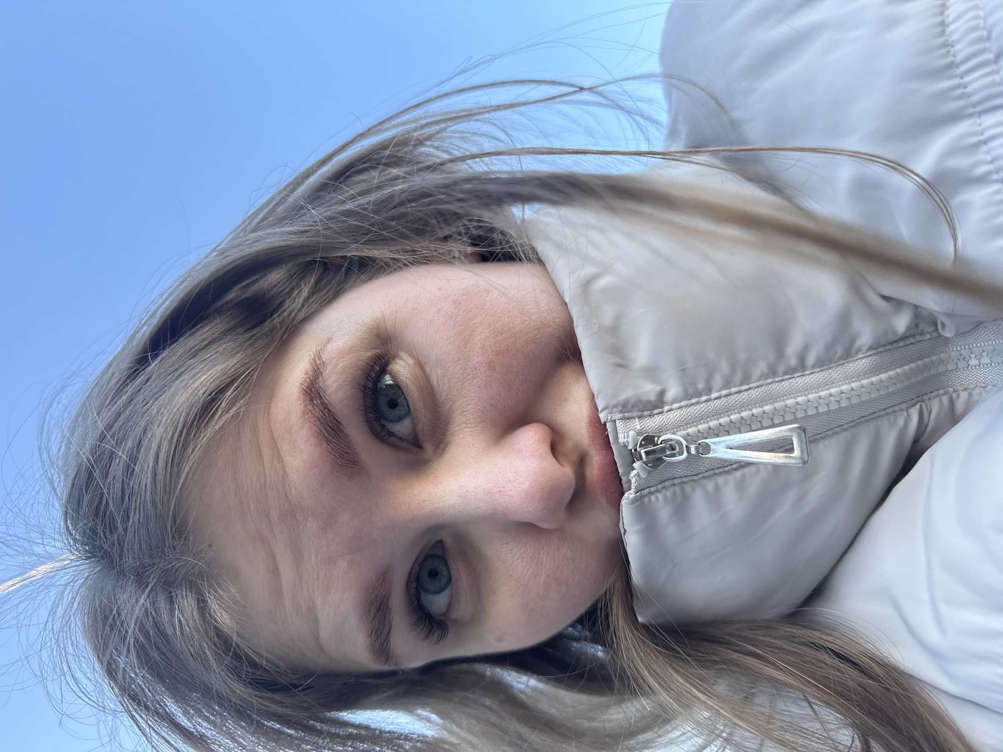 A close-up, angled portrait shows a fair-skinned person with blue eyes and light brown/grayish hair. They are wearing a puffy white jacket with a visible silver zipper. The background is a clear blue sky. The location is Горный, Russia.