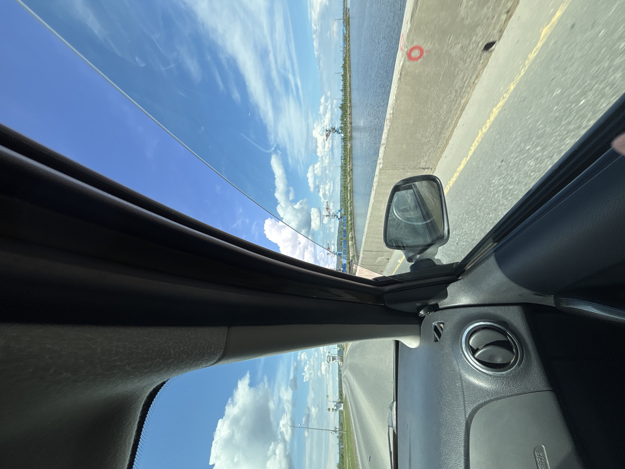 The image is taken from inside a car, looking out and slightly upward through the open sunroof. The car is on a bridge, next to a body of water. The sky is visible through the sunroof and side window, featuring blue sky with scattered white clouds. The car's interior is visible, including the side mirror, dashboard, and air vent. The bridge has a concrete barrier with a red circular marking. The landscape beyond the water includes greenery, industrial cranes, and power lines. The road surface has a yellow line marking. I cannot confirm the location as Chaikovsky, Russia, based on the visual information provided.