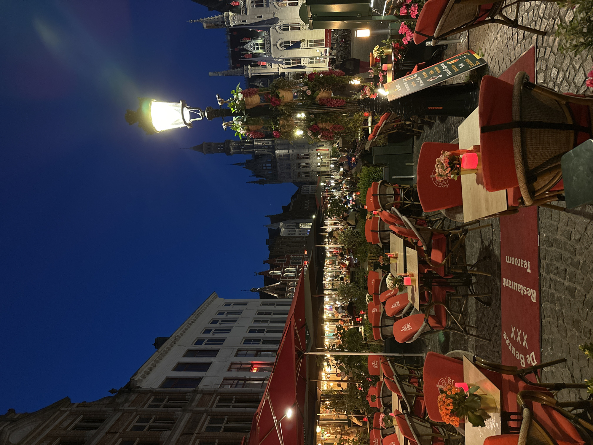 The image depicts an outdoor seating area of a restaurant or tearoom in Bruges, Belgium, at dusk. The scene is characterized by an array of red chairs and tables set on a cobblestone ground. Red umbrellas provide shade over some of the tables. Flower arrangements in vases and hanging baskets adorn the area, adding pops of color. A lamppost with ornate floral decorations stands prominently in the foreground, casting a warm glow. In the background, illuminated buildings with traditional Flemish architecture are visible, their facades lit against the darkening blue sky. Several people are seated at tables in the distance, engaged in conversation or dining. A red mat with the text "De Beuze XXX Restaurant Tearoom" is placed in front of some of the tables. A sign board stands near the lamppost, displaying menu items. The overall atmosphere is that of a lively yet relaxed evening setting in a historic European city.