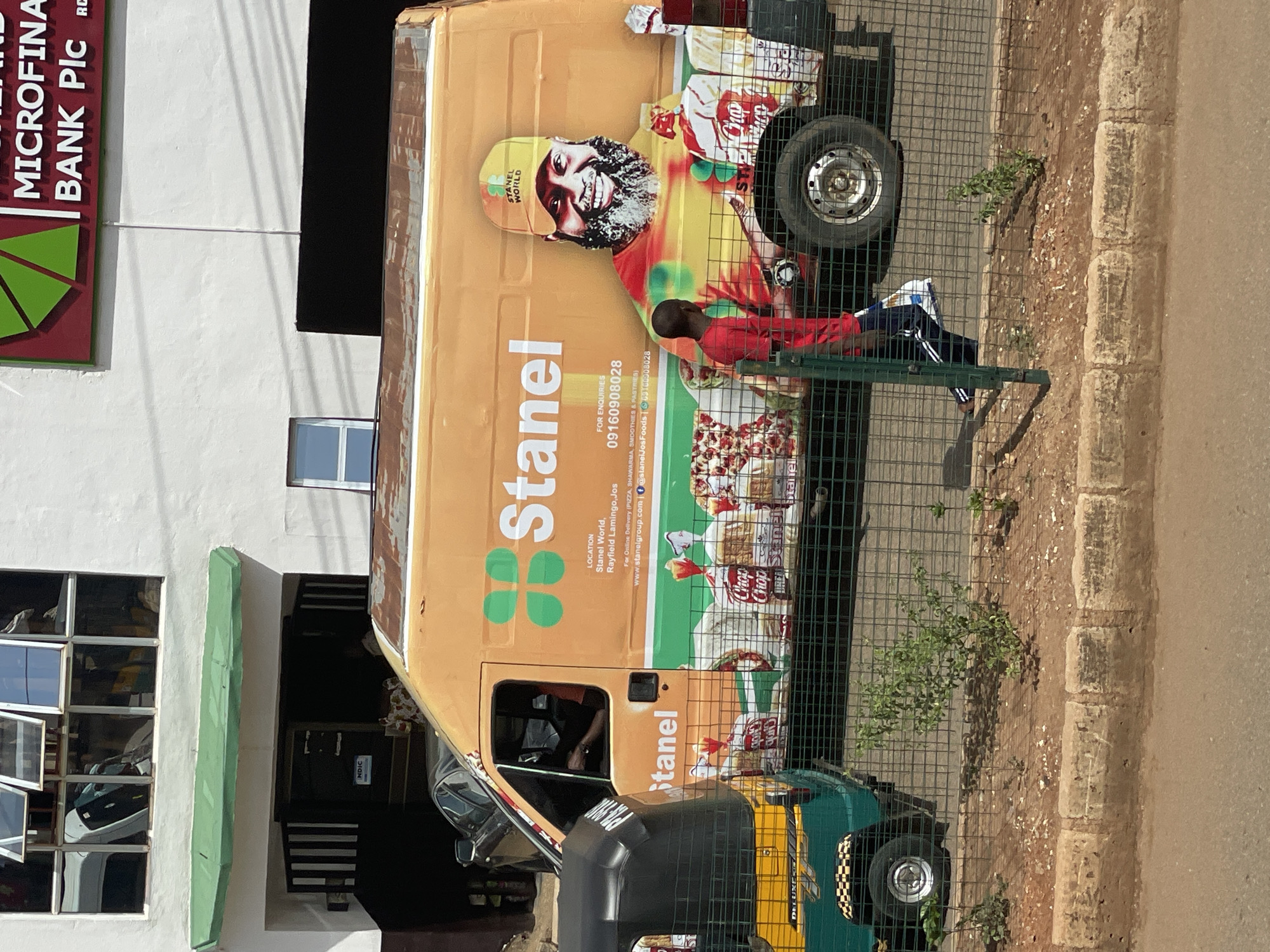 The image depicts a Stanel World delivery van parked beside a building, partially obscured by a wire fence. The van is painted with an advertisement featuring an individual wearing a Stanel World branded cap and promoting food items like pizza and "Chop Chop" branded products. A young person wearing a red shirt and blue trousers stands close to the van, on the side of the street. The location is likely in Bukuru, Nigeria, based on the "Stanel World" business indicated on the van, which has a location listed in Rayfield Lamingo, Jos. A building with a sign for a microfinance bank is partially visible in the background. The surrounding environment includes a sidewalk and a road.