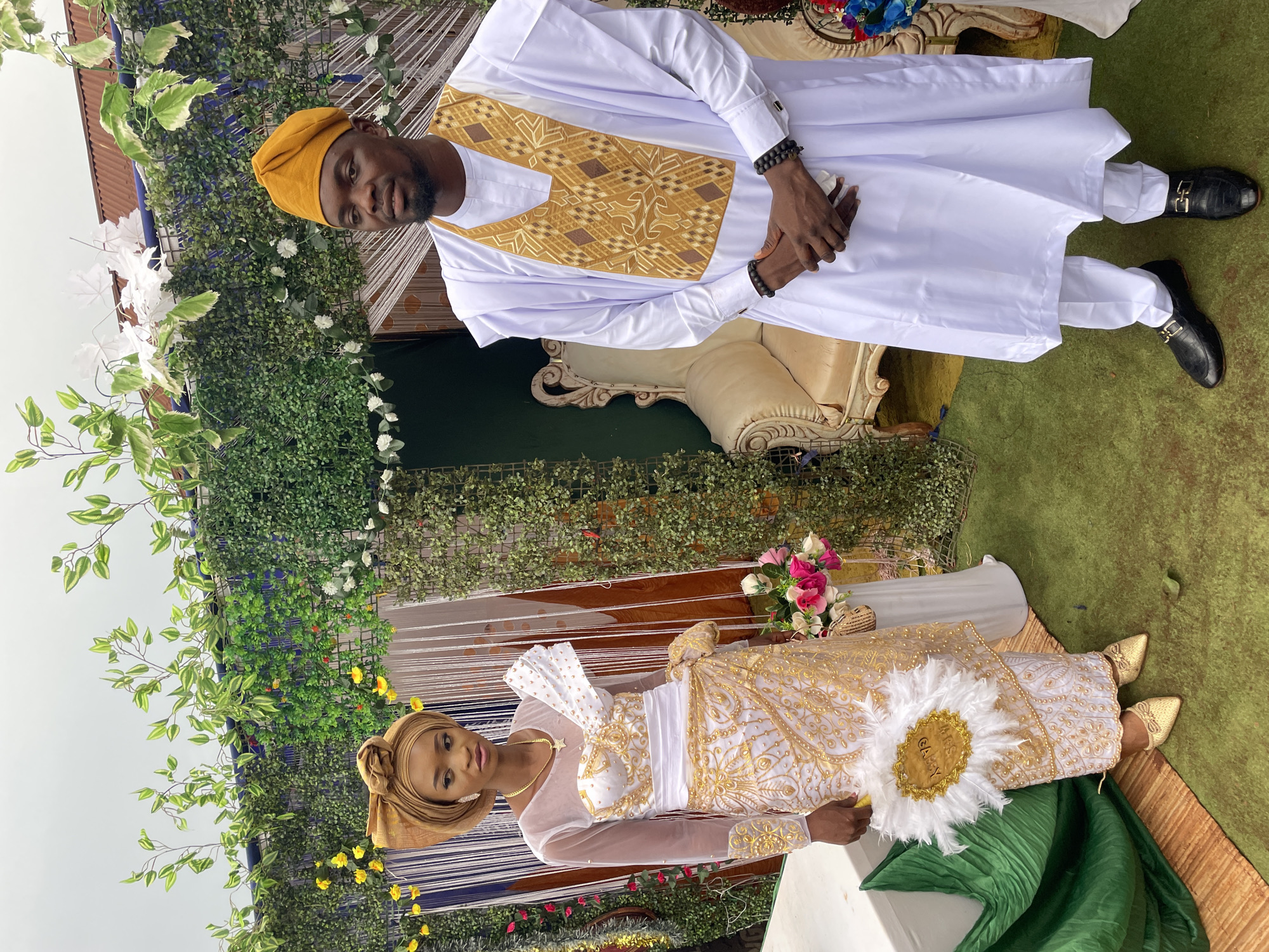 A couple poses for a photo in what appears to be an outdoor event setting in Ogbe, Nigeria. The man stands behind a cream-colored couch, wearing a white traditional outfit with gold embroidered details on the chest, a matching yellow cap, and black shoes. He has dark skin and is wearing beaded bracelets. The woman is seated on the ground, wearing a white and gold traditional dress with a matching headwrap, gold shoes, and jewelry. She holds a white feathered fan with gold detailing. The background includes greenery, flowers, and what seems to be decorative netting. The sky is overcast.
