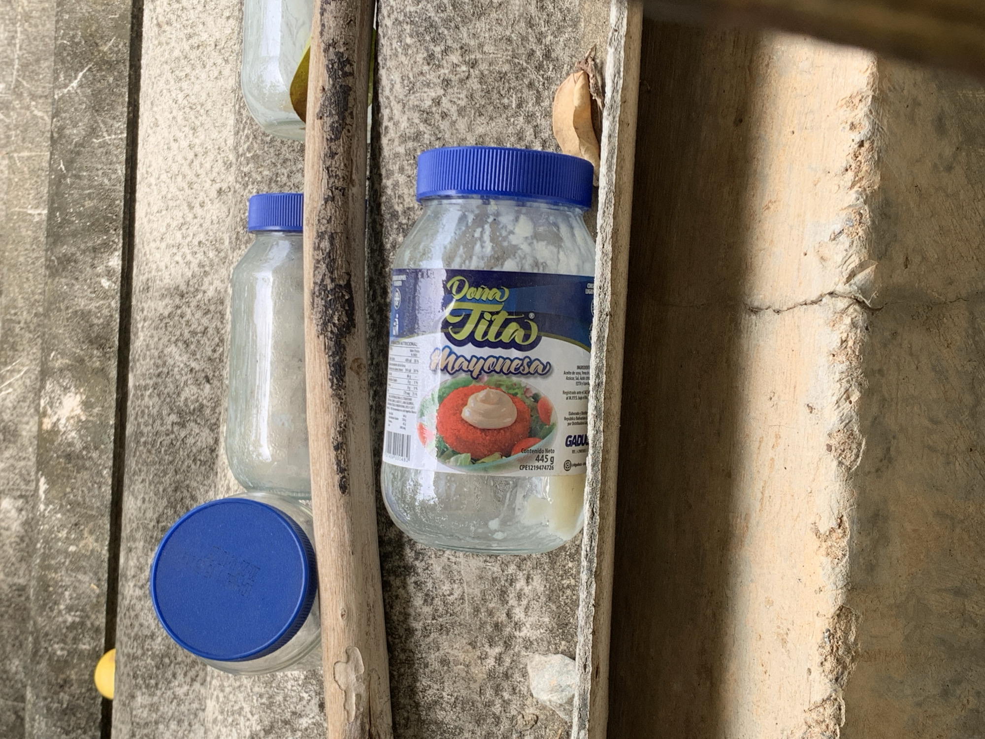 The image depicts a static arrangement of discarded or temporarily placed objects on a series of worn concrete steps, located in Santa Ana, Venezuela. In the foreground, a clear glass jar, identified by its label as "Mayonesa Pura Tita" (Net Content 445 g, CFE131907026), lies on its side with a blue screw-top lid and a small amount of light-colored residue inside. The label features an image of a red, spherical food item with a dollop of mayonnaise. A dry, brown leaf rests near the jar's lid. To the left of this jar, a small, pale stone is visible on the concrete step. A rough, natural wooden stick with visible bark runs horizontally across the mid-ground, positioned beneath the glass containers. Further back and to the left, another clear glass jar with a blue lid is oriented horizontally on an upper step. In the mid-ground, two additional clear glass bottles are visible, also lying on their sides; one has a blue screw cap, while the cap of the other is not clearly discernible. A yellow object is partially visible on a higher step in the upper left background. The concrete steps are textured, exhibit signs of wear, and show variations in color consistent with outdoor exposure and accumulated dirt. No individuals are present in the scene.