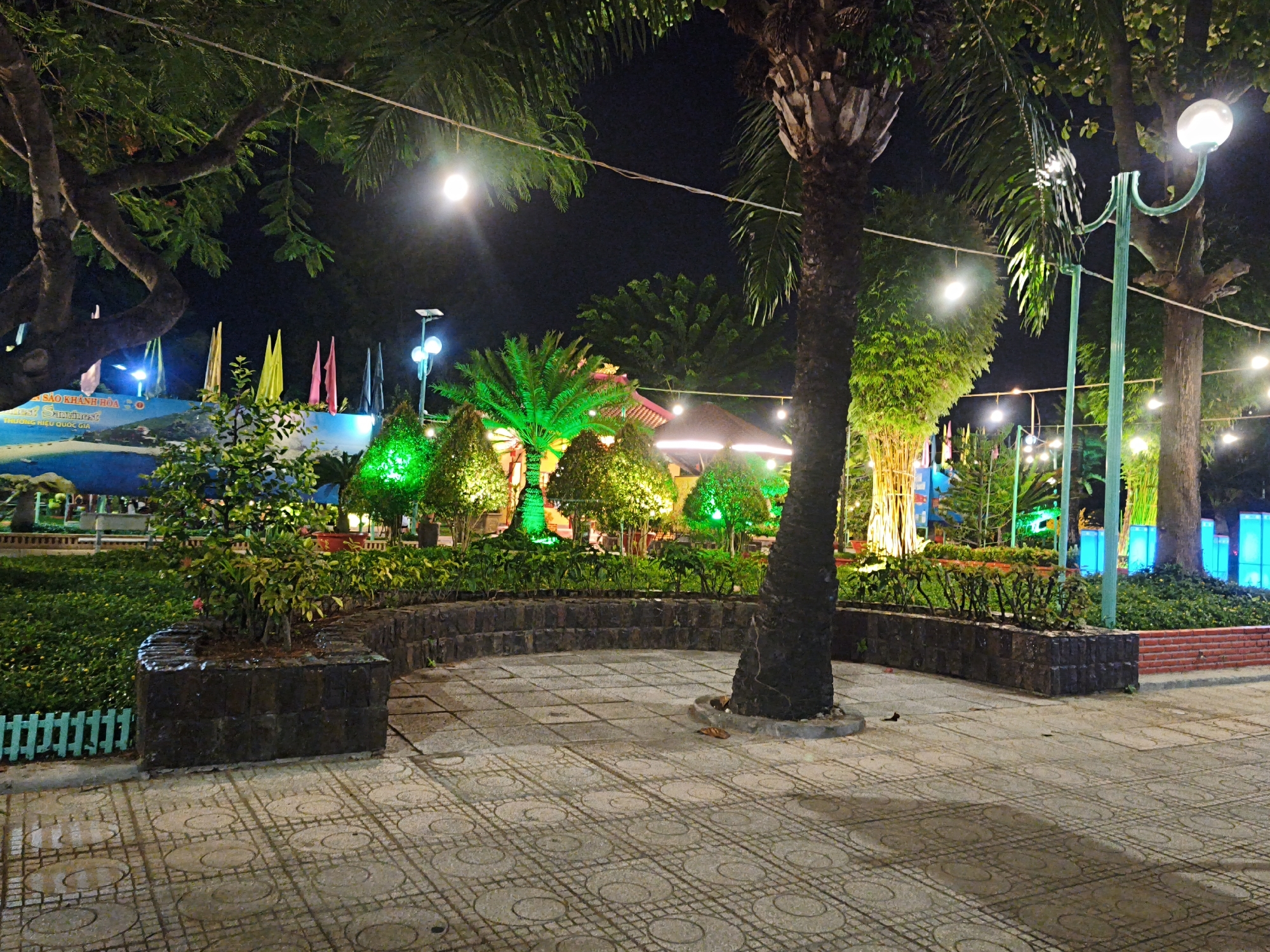 The image depicts a park scene at night in Bac Nha Trang Ward, Vietnam. The foreground is dominated by patterned paving stones, some of which appear wet. A large, dark planter box sits to the left, containing a small tree with green and yellow leaves, some with reddish blossoms. A low, light green picket fence runs along the edge of the planter. Behind this, a curved stone wall encloses a garden area with lush green foliage.
In the midground, several palm trees and other verdant plants are illuminated with vibrant green lights. A dark-colored, tiered structure, possibly a gazebo or small building, is visible in the background, with glowing lights. Strings of small, yellow lights are strung across the scene, and a decorative streetlamp with a curved green metal arm and a white globe is present on the right. Beyond the garden, a blue and white mural or backdrop is visible, adorned with colorful flags in yellow, pink, and black.
The background features more trees and a dark night sky. The overall impression is of a well-lit, festive park environment at night.