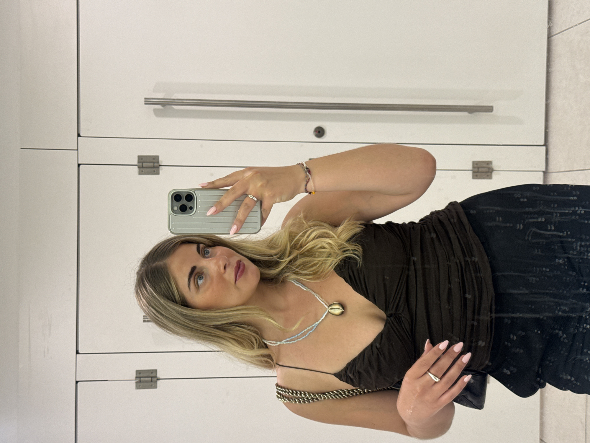 A woman takes a selfie in a public restroom, likely located in Paris, France. She is holding a light green iPhone with a ridged case in her left hand, her arm bent at the elbow, with her right hand also in view. She is wearing a brown, sleeveless top with spaghetti straps and a black, patterned skirt. She has a necklace featuring white, blue, and gold beads. She has blond hair and is looking at the camera. The background is a wall of white lockers with brushed steel handles.