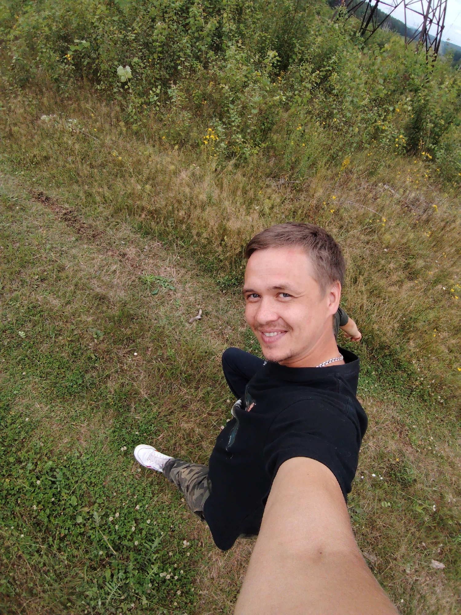 The image shows a man taking a selfie outdoors. He is wearing a black hoodie, camouflage pants, and white sneakers. He is smiling and looking at the camera. The background is a grassy field with some bushes and trees. There are also some power lines visible in the distance. The image appears to have been taken during the daytime. The location in the city of Прикамский, Russia is not provided in the image.