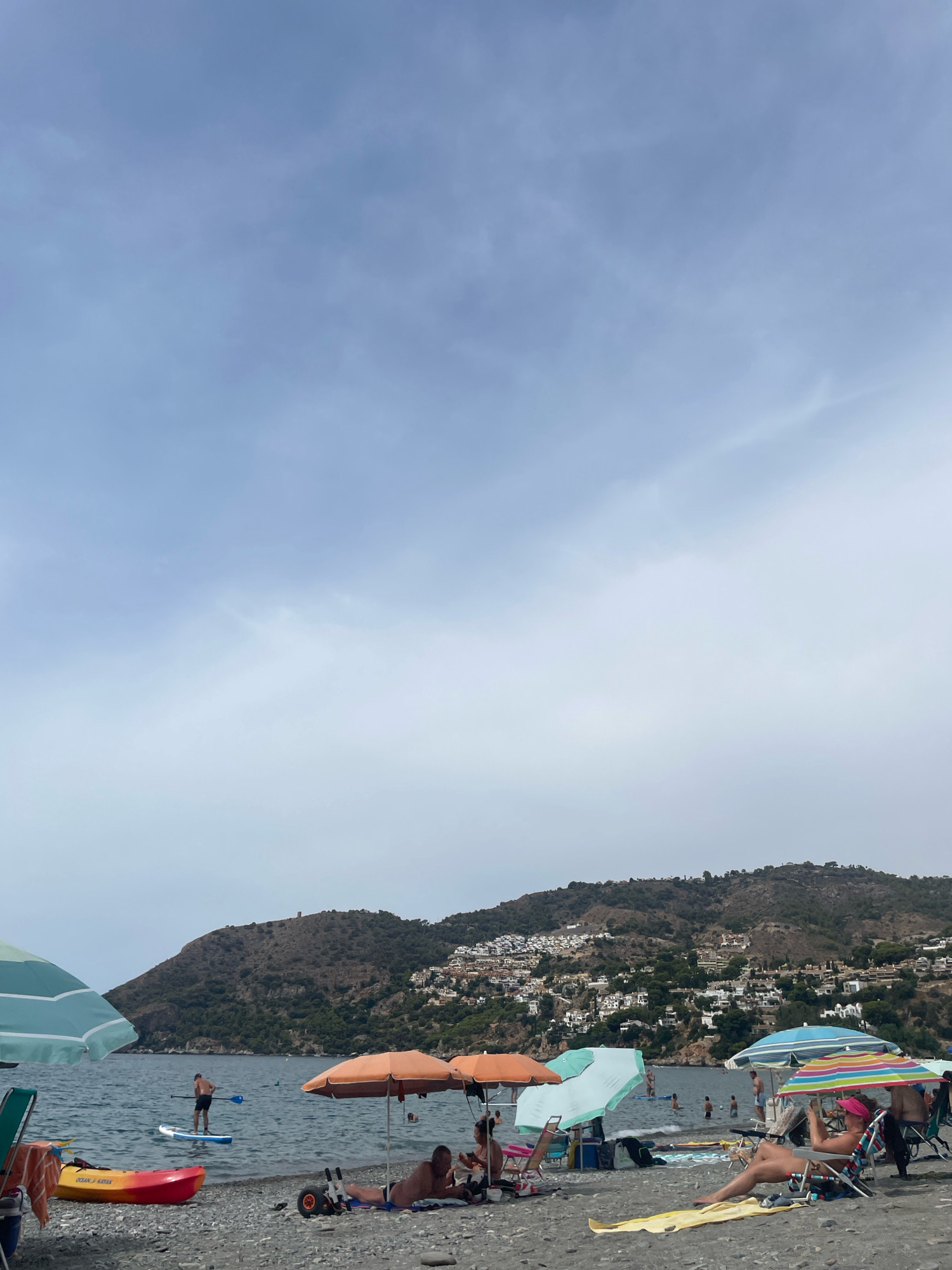 The image depicts a beach scene in La Herradura, Spain. The foreground is occupied by a pebble beach with several beach umbrellas providing shade. People are seated or reclining on beach chairs under the umbrellas, and one person is lying on a yellow towel. A kayak is visible on the left side of the beach. In the water, a man is paddleboarding, and several other people are swimming. Beyond the water, a lush, green hillside is covered with a white village, characteristic of the coastal architecture of the region. The sky is overcast with soft clouds.