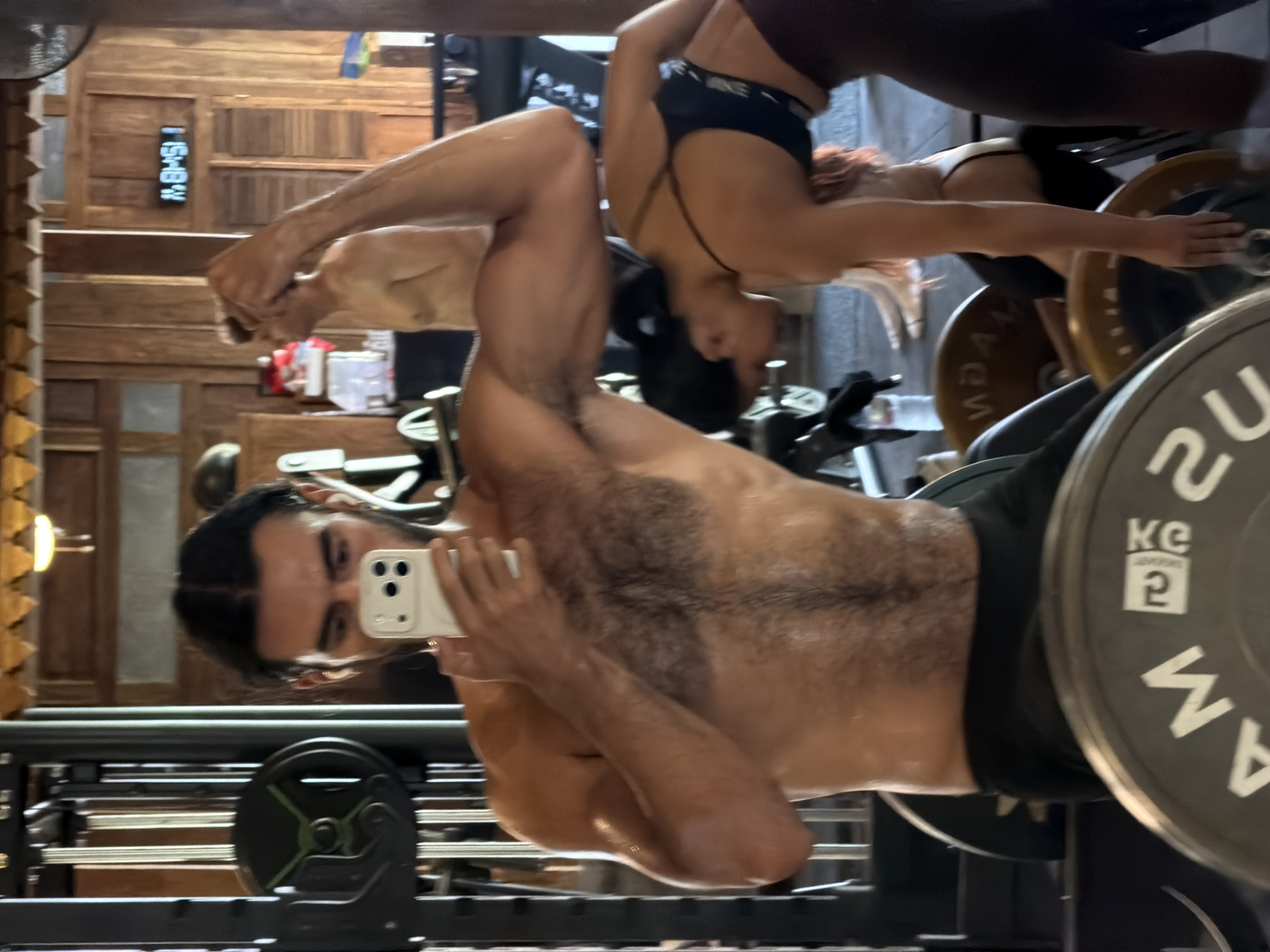 A mirrored image depicts two individuals in what appears to be a gym in North Kuta, Indonesia. One person, shirtless, stands close to the mirror, holding a smartphone and seemingly taking a picture of himself. He is muscular and has dark hair and a beard. The second person, visible in the mirror's reflection, is wearing activewear and is engaged in some form of exercise involving weights. Gym equipment, including a barbell with weights, is visible in the foreground. The background reveals wood paneled walls and a digital clock displaying time.