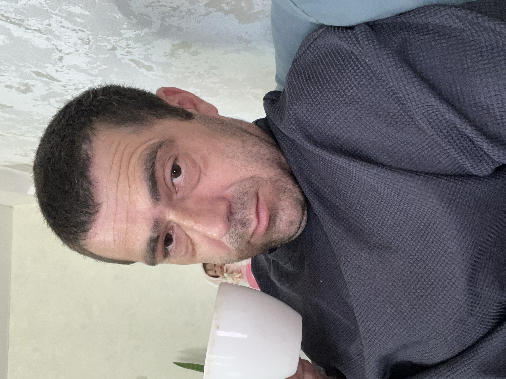 A man is holding a white coffee cup in his left hand, and looking at the camera. He is wearing a dark gray, textured shirt. In the background, a child wearing a pink outfit is visible. The setting appears to be indoors, with a light-colored wall and a textured wall behind the man. There is also a glimpse of a blue couch. The overall lighting suggests daytime.