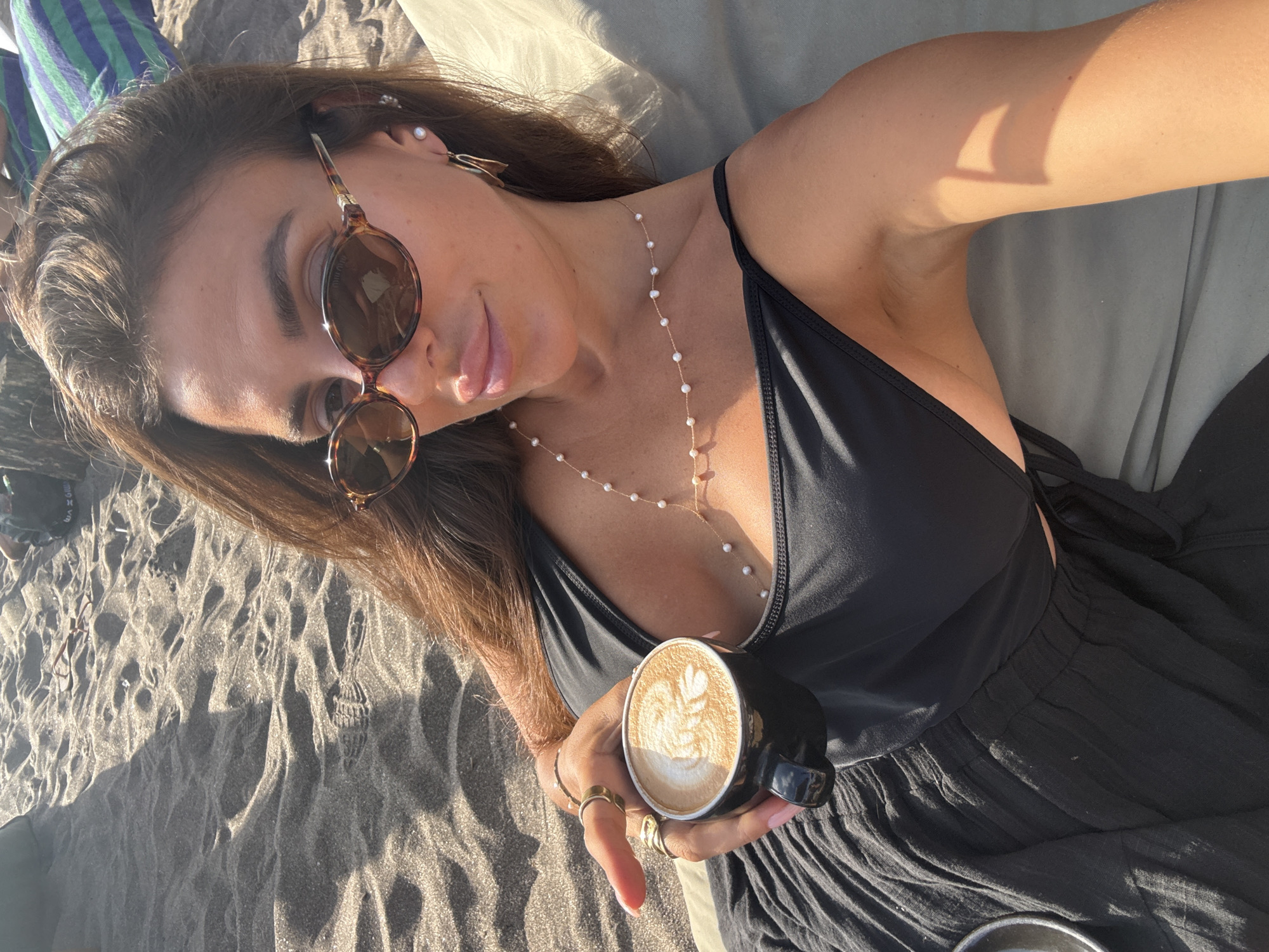 A female subject is depicted reclining on a light-colored lounge cushion at a beach in North Kuta, Indonesia. She has long, dark brown hair and wears tortoiseshell oval sunglasses, a black spaghetti-strap top, and a long, dark, textured skirt. Her left hand, adorned with multiple gold-toned rings and light pink nail polish, holds a black cup containing a latte with a discernible leaf-patterned art design. A delicate gold-toned chain necklace with small pearls is worn around her neck. Her right ear features a pearl stud earring and a larger gold-toned abstract hoop earring. The background consists of dark-grained sand with visible footprints and ripples, illuminated by direct sunlight which casts sharp shadows. Indications of other human presence and a striped fabric item are partially visible in the distant background on the sand.