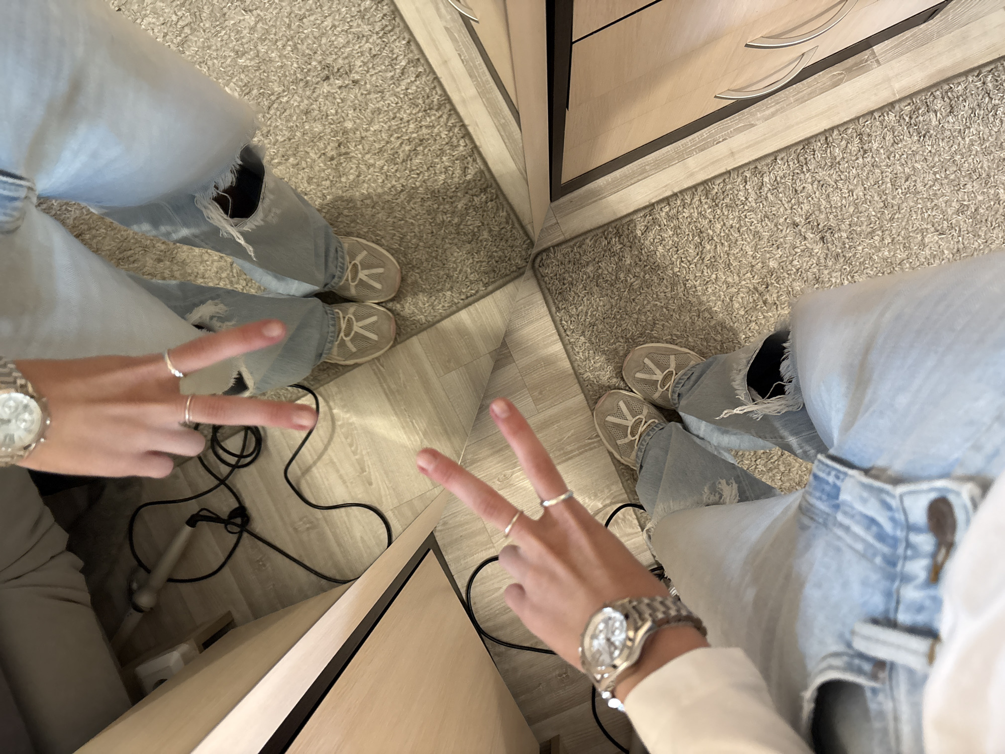 The image shows a close-up view of two people's legs and hands, as seen from a high angle. Both individuals are wearing light-wash ripped jeans and athletic shoes. One person is wearing a silver watch on their left wrist and is holding up two fingers in a peace sign with their left hand, adorned with two silver rings. Their right hand, also with two rings, is visible near their jeans. The other person's hands are not fully visible.
The environment appears to be indoors, possibly a room with a light-colored carpet and wood-paneled furniture or walls. A black cord from a hair styling tool is visible on the floor. The flooring is a mix of wood laminate and carpet. Reflections in a mirror or reflective surface are also present, showing parts of the scene from a different perspective. There are no elements in the image that would suggest a location in the city of Perm, Russia.