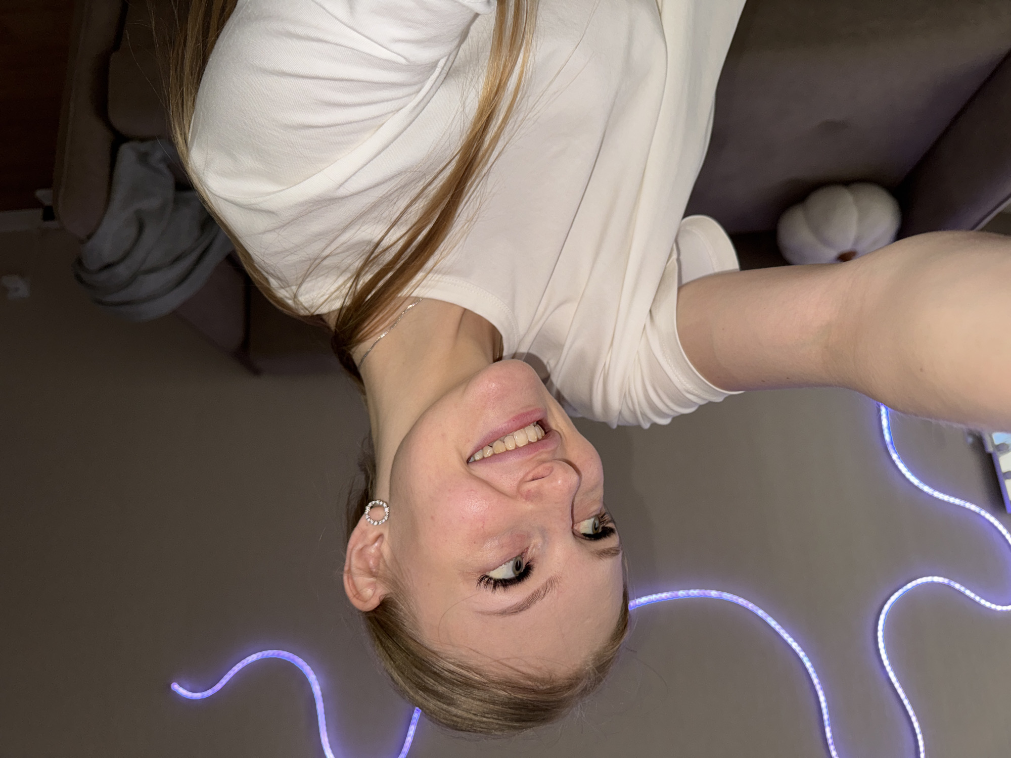 A young woman with blonde hair is captured in a selfie, her head tilted to her left. She's wearing a white t-shirt and a circular, jeweled earring in her left ear. Her eyes are open, and she has a wide smile, revealing her teeth. The background is dimly lit, with a faint purple glow from what appears to be LED strip lighting. To the right, a white, gourd-shaped object is visible on a dark surface. The image seems to be taken indoors, possibly in a residential setting. There is no information in the image to suggest a location in the city of Perm, Russia.