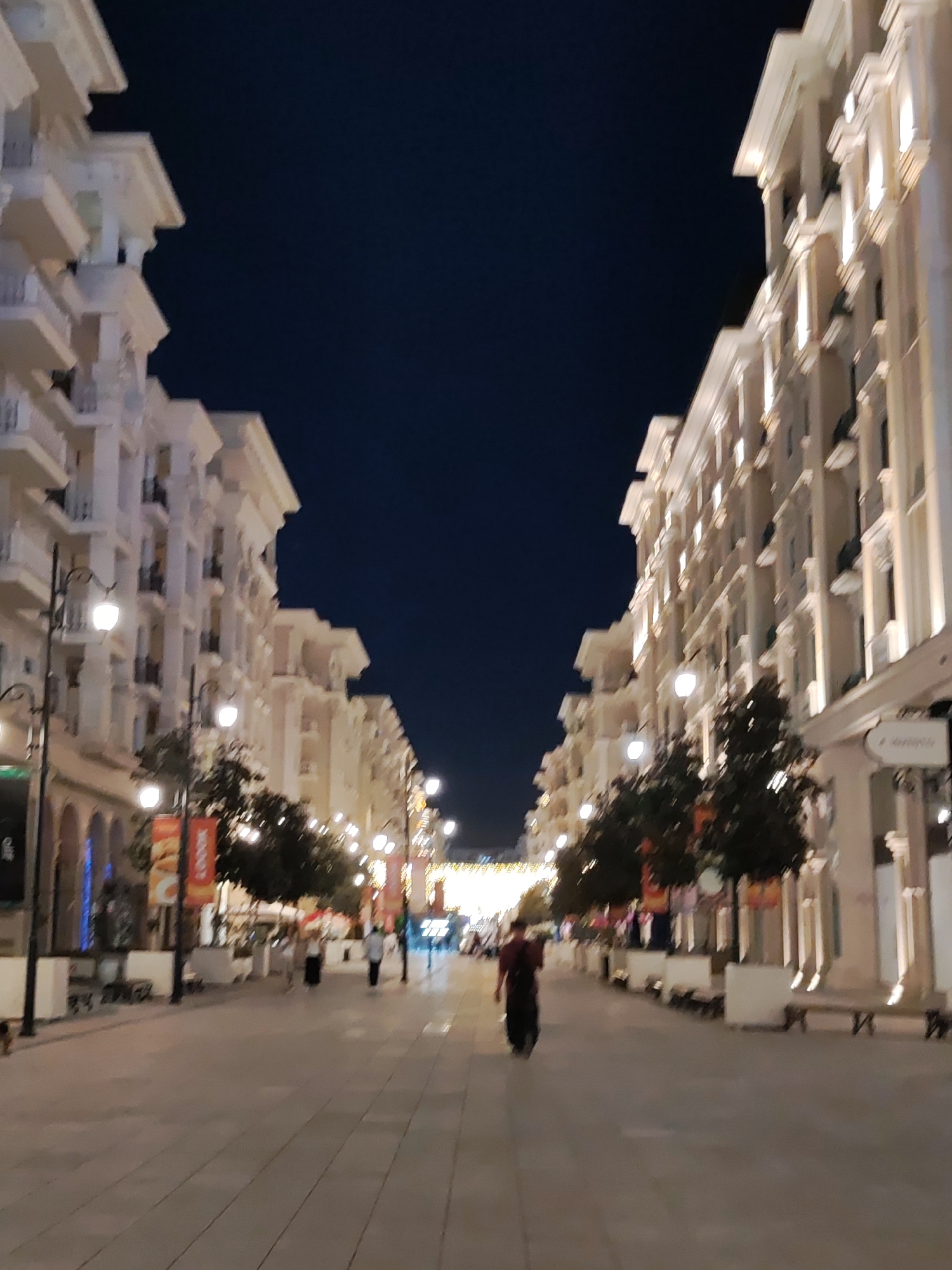This is a vertical, slightly blurry, night-time photograph of a pedestrian street lined with white, multi-story buildings on both sides. The buildings have decorative architectural details, including balconies and columns, and are illuminated by warm lighting. The street is paved with large, light-colored tiles. Several streetlights with ornate metal posts and glowing lamps are visible along the sidewalk. Trees with dark foliage are planted along the sides of the street. There are a few people walking on the street. In the foreground, a person in a dark shirt and pants is walking away from the camera. Further down the street, more figures are visible, along with some businesses with illuminated signage. The sky is a deep, dark blue. The scene appears to be in the city of Tashkent, Uzbekistan, likely a newly developed area or a well-maintained historic street given the architecture.