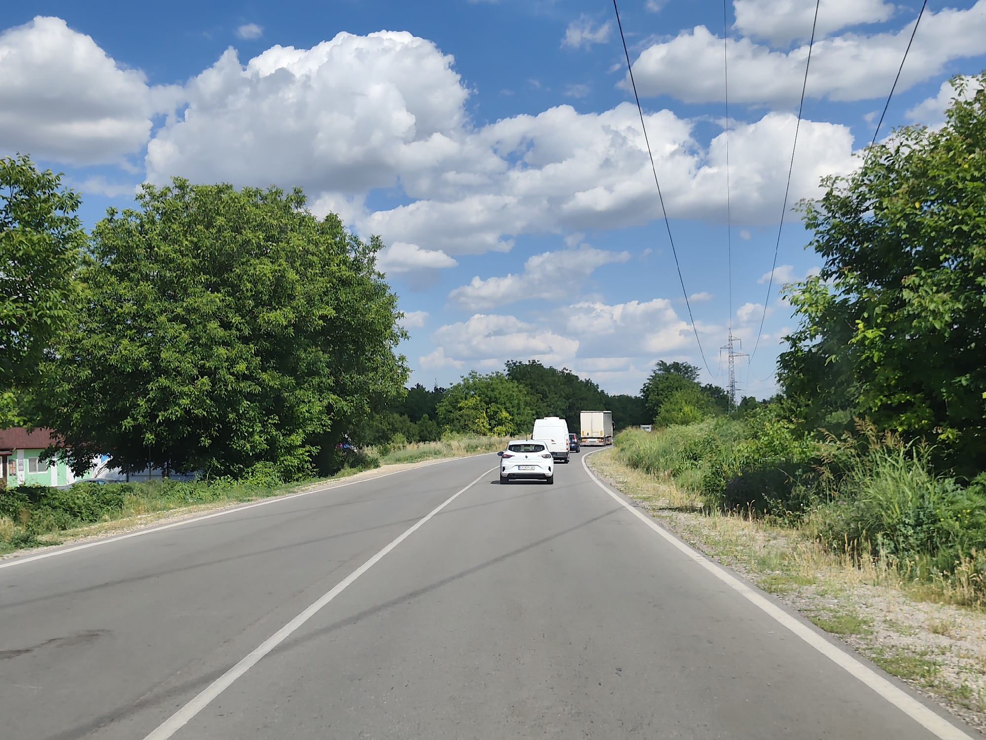 This is a landscape, eye-level, medium shot of a two-lane asphalt road curving slightly to the right. The road is bordered by a white dashed line on the left and a solid white line on the right. A white passenger car is in the right lane, followed by two white panel vans and a large semi-truck with a trailer in the distance. The road is lined with lush green trees and vegetation on both sides. The sky is a vibrant blue with fluffy white clouds, and power lines stretch across the frame. In the far distance on the left, a house with green trim is visible. The scene is likely located on a rural road outside of Pleven, Bulgaria, given the typical appearance of rural infrastructure and vegetation in the region. The overall impression is a clear, sunny day with moderate traffic.
