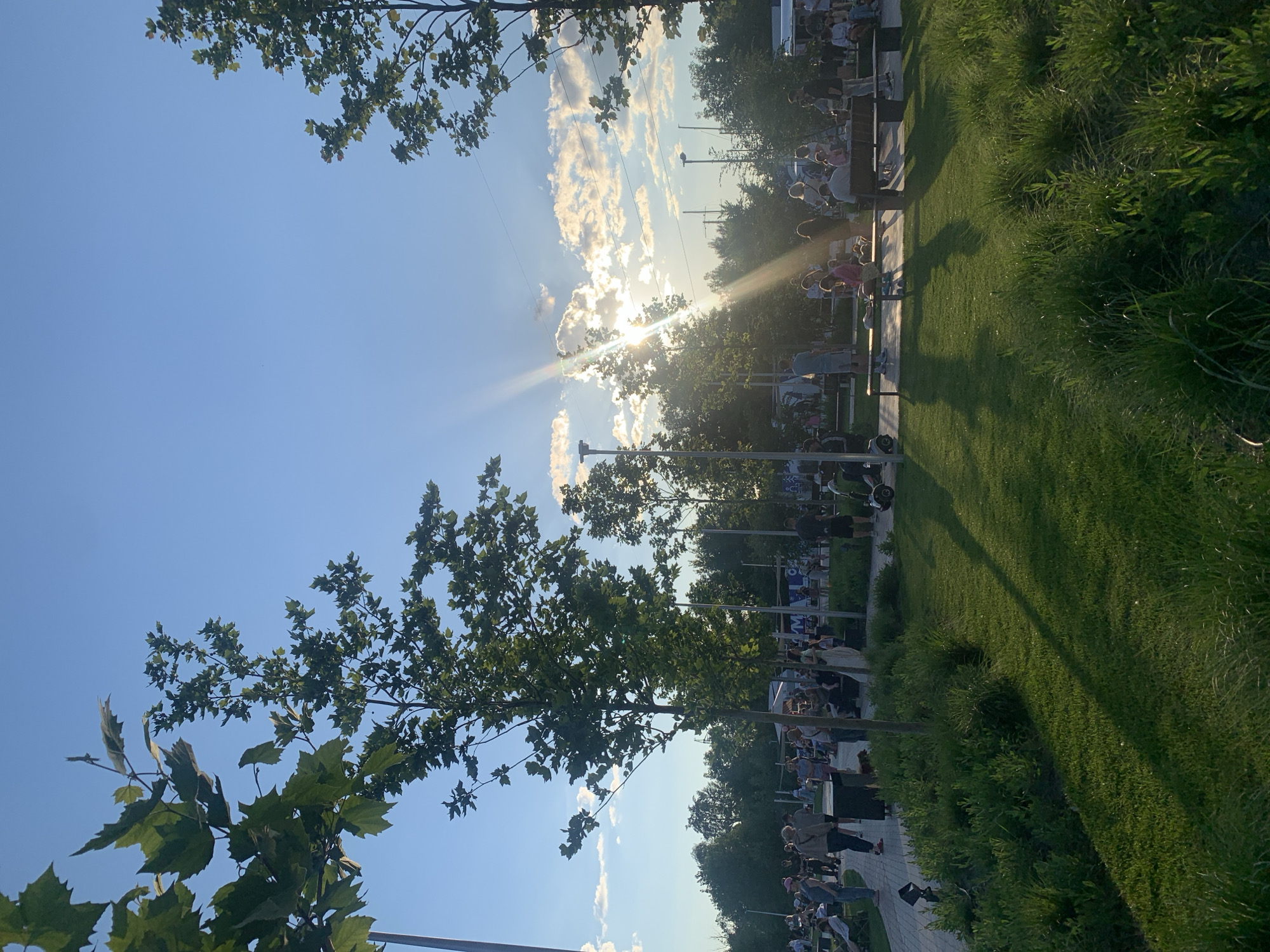 The image depicts a scene in a park with people gathered in the late afternoon or early evening. The sun is low in the sky, casting long shadows across the green grass and casting a bright lens flare through the trees. In the foreground, lush green grass and bushes dominate, with a paved walkway visible to the left. Several trees with green foliage are scattered throughout the park, some in the foreground and others further back.
In the middle ground and background, a gathering of people can be observed. Some are walking along the pathway, while others are sitting on benches or standing in small groups. There appear to be some structures or stalls set up, and various individuals are engaged in recreational activities. One person is seen on a hoverboard or Segway. The overall atmosphere suggests a public event or a relaxed outing in a park setting. The scene is characterized by the interplay of sunlight, shadows, and the natural elements of the park.
Based on the context provided in the prompt, this scene is located in the city of Kyiv, Ukraine.