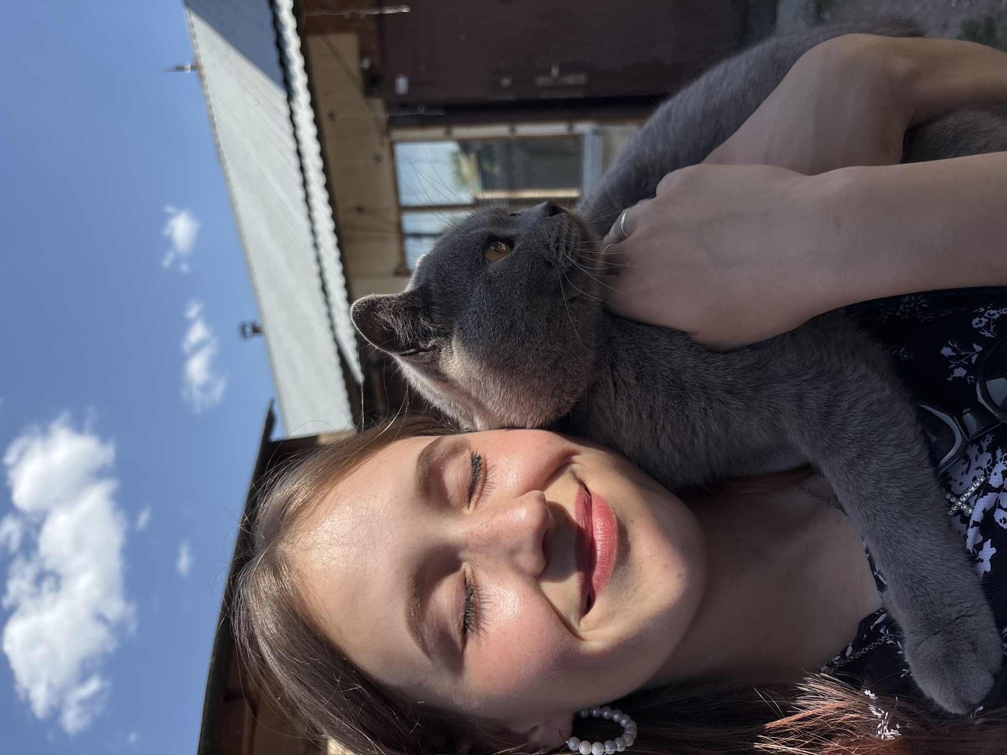 A young woman lies on her back, eyes closed and smiling, with a gray cat resting on her chest and shoulder. The cat is looking upwards and to the left. The woman is wearing earrings and a patterned top, and someone's hand is resting on the cat. Above and behind them is the blurred background of a building and a bright blue sky with white clouds. The location is Фоки, Russia.