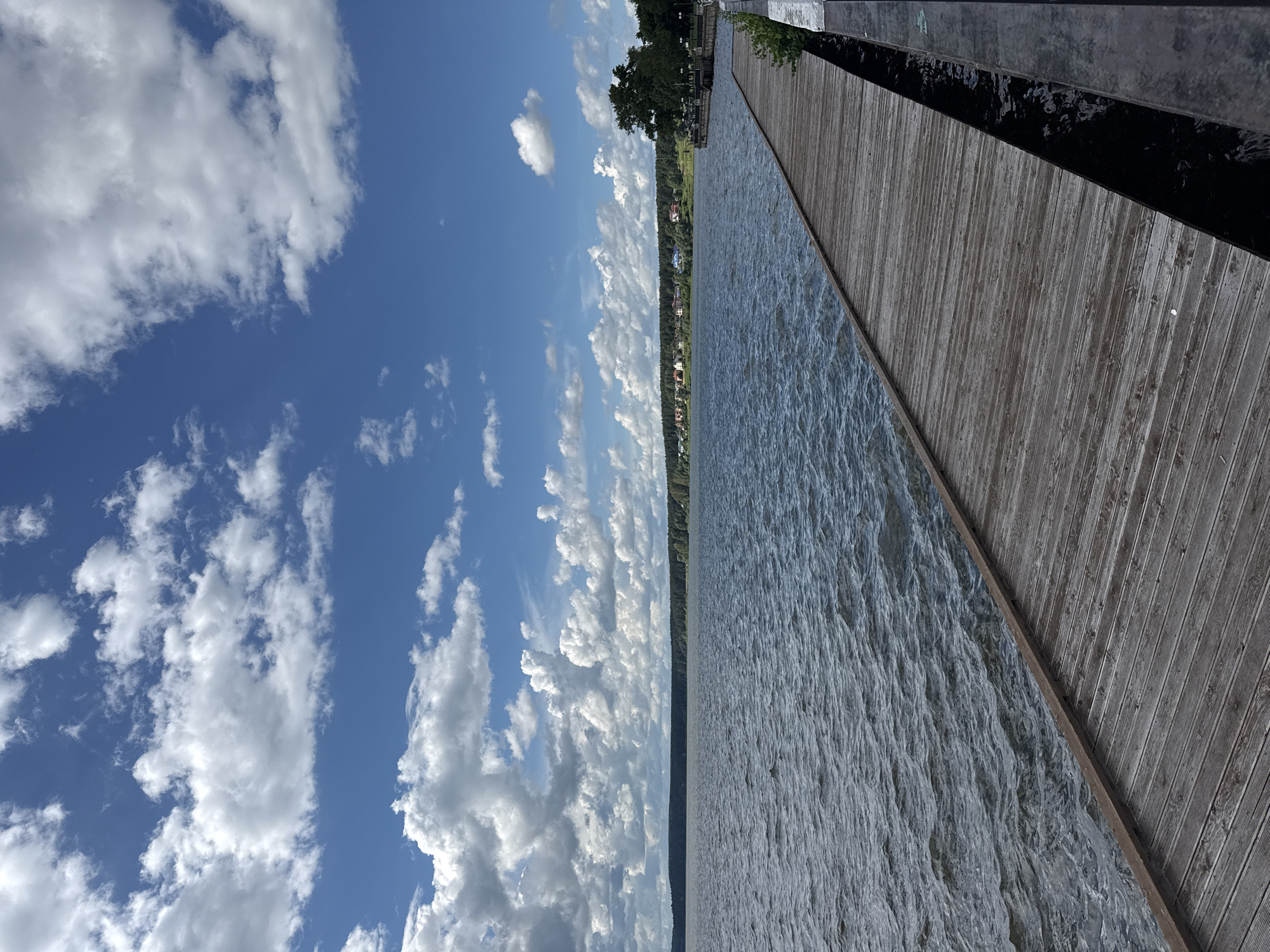 An outdoor vertical, eye-level shot captures a wooden boardwalk extending into a large body of water. The boardwalk, constructed from weathered wooden planks, occupies the lower right portion of the frame, angling away from the viewer. To the left of the boardwalk is a vast expanse of dark, rippling water, with small waves visible. The horizon line is established by a dense, dark green treeline that stretches across the mid-ground, punctuated by scattered buildings of various colors, indicating a settlement. Above the treeline, a bright blue sky is filled with dramatic, white cumulus clouds that cast shadows, suggesting a dynamic weather system. The overall impression is one of a serene, natural landscape on a partly cloudy day. This scene is likely the embankment along the Votkinsk Reservoir in Votkinsk, Russia, a popular recreational area.