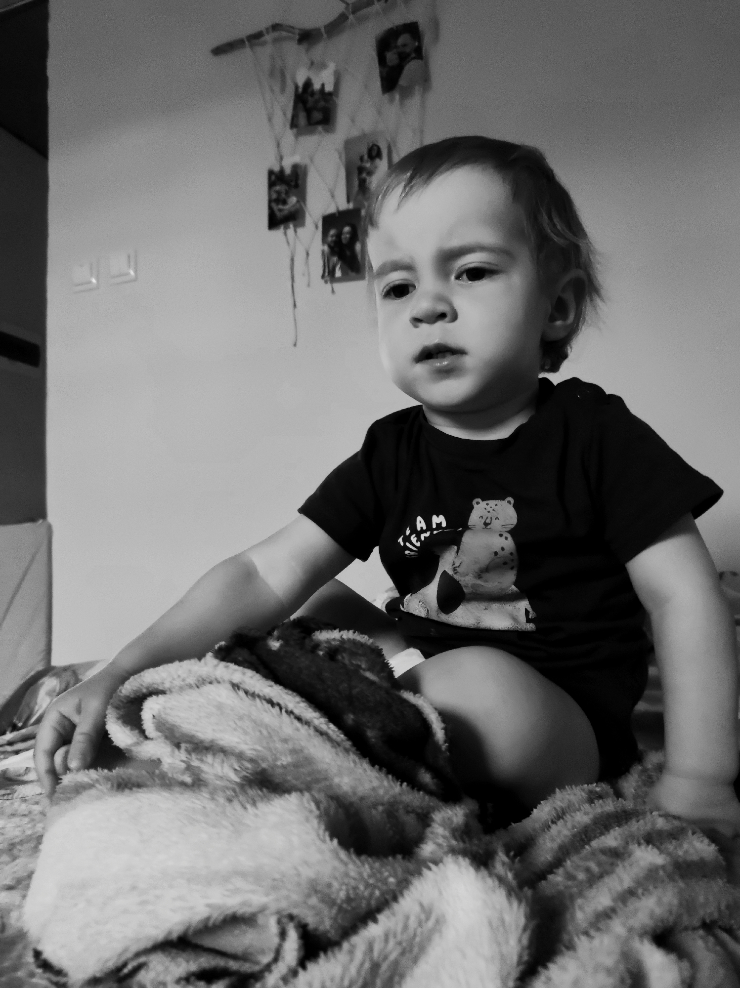 A black and white photograph captures a young child, likely a toddler, seated on a bed or soft surface covered by a textured, fluffy blanket. The child has light-colored, medium-length hair and a round face, looking towards the right side of the frame with a serious or pensive expression, mouth slightly ajar. They are wearing a dark short-sleeved t-shirt featuring a graphic of a spotted animal, resembling a cheetah or leopard, and partially visible text that reads "TEAM" with another word below it, possibly "TEN," which is partially obscured. One of the child's hands rests on the blanket.
The background reveals an indoor setting, likely a residential bedroom. A light-colored wall displays a decorative hanging made from a branch or stick with strings from which several small framed photographs are suspended. To the left of the child, two light-colored rectangular fixtures, possibly light switches or outlets, are visible on the wall. A darker vertical structure, possibly a door frame or furniture, is partially visible on the far left edge of the image. The monochrome presentation gives the image a classic, timeless feel. The time of day is not discernible, and no specific event beyond the child sitting and observing is depicted.