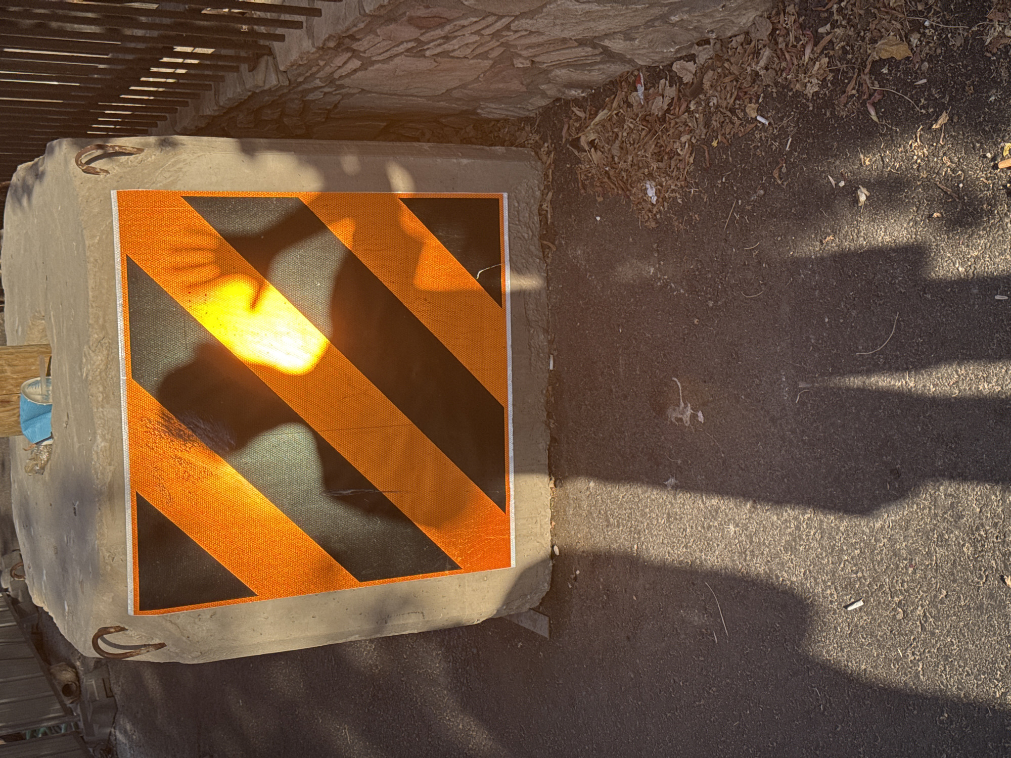 A concrete barrier with a hazard sticker featuring orange and black diagonal stripes is positioned on an asphalt surface. A shadow of a hand with outstretched fingers is cast onto the sticker, illuminated by a bright light source, possibly sunlight. The scene is outdoors, with a stone wall and debris, including dry leaves and cigarette butts, visible in the background. The lighting suggests it is daytime, likely late afternoon or early morning due to the long shadows. The overall impression is one of an urban or semi-urban environment in Tel-Aviv, Israel, with the barrier likely serving as a safety or traffic control measure.