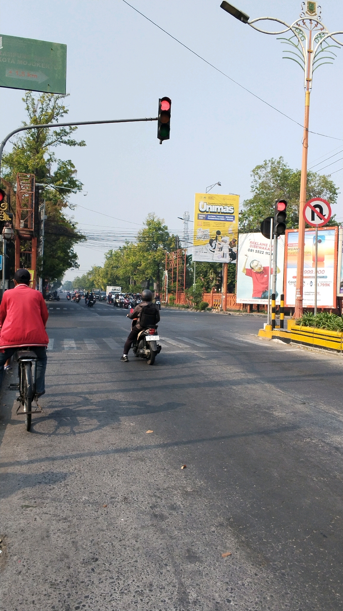 In Kranggan, Indonesia, a busy street scene unfolds under a clear sky. A person on a bicycle, viewed from behind, wears a red jacket and blue jeans, facing away from the camera on the left side of the road. Ahead of them, a motorcyclist in a dark helmet and jacket, also viewed from behind, rides a black scooter. Both vehicles are positioned on the right side of the road, with the traffic light overhead showing red. The road is asphalt, with a pedestrian crossing marked in white lines. On the right side of the road, a tall traffic signal pole stands with a red light illuminated. Beside it, a large billboard displays advertisements for "Unimas" and "Huawei". Behind the billboard, trees and other structures are visible. The overall environment suggests an urban setting with moderate traffic.