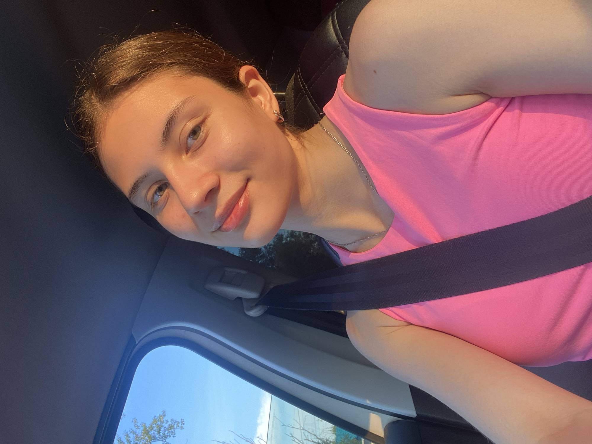 A young woman with light brown hair and light skin is in the passenger seat of a car. She is wearing a bright pink tank top and a silver chain necklace. She is looking at the camera with a slight smile and her head is tilted to her left. The sun is shining on her face, creating a warm glow. A seatbelt is visible across her chest. The background shows the car's interior and a blue sky with some trees visible through the window. There is no discernible location in Votkinsk, Russia.