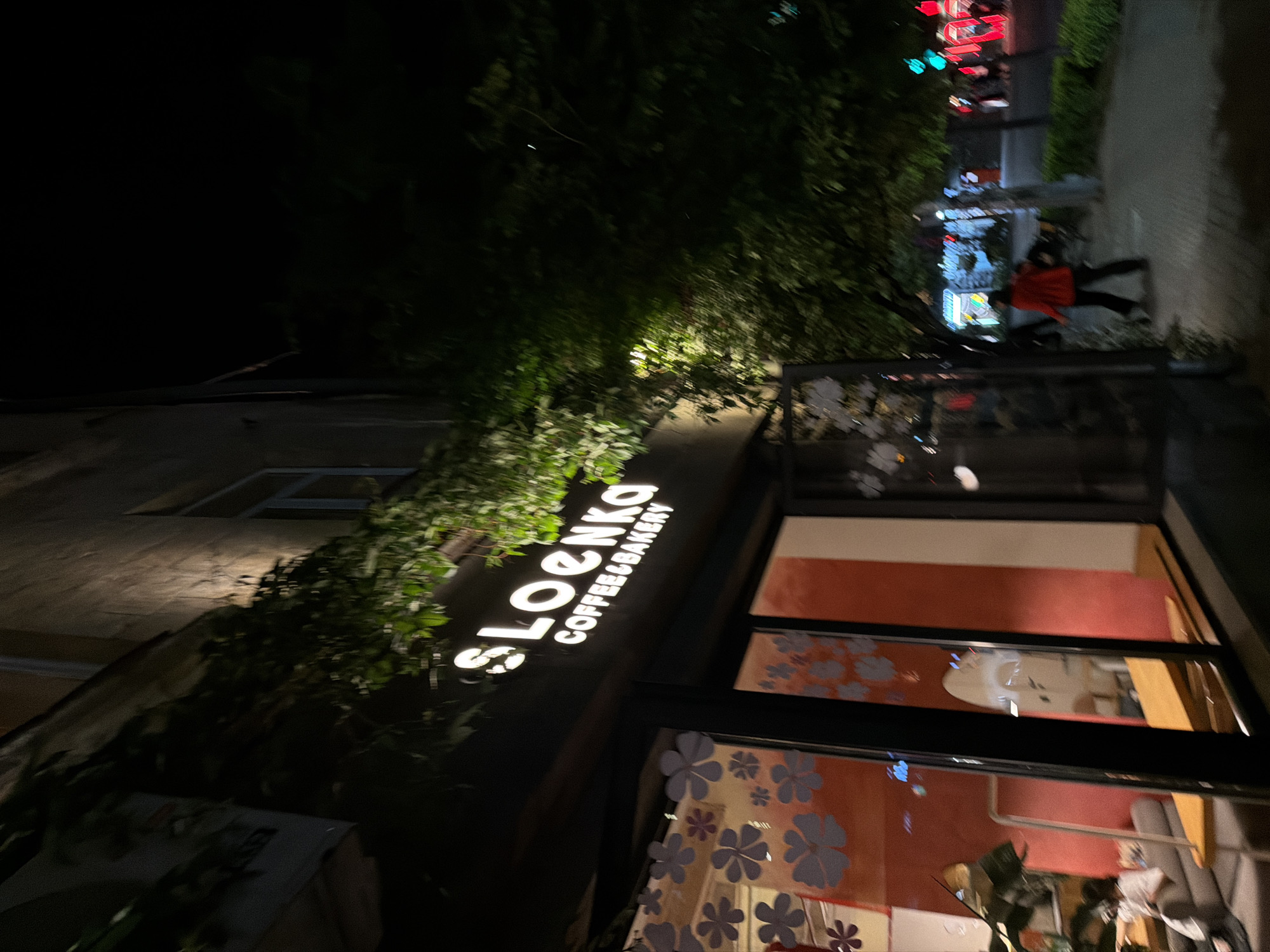 The image depicts the exterior of "SLoenka Coffee&Bakery," a business in Yerevan, Armenia, at night. The building is partly obscured by foliage. The illuminated sign of the coffee shop is visible above the storefront. Large window panes display decorations of flower patterns. Red and white interior walls are seen through the windows. A person wearing a red jacket walks along the sidewalk in the distance. Streetlights and signs are also visible in the background.