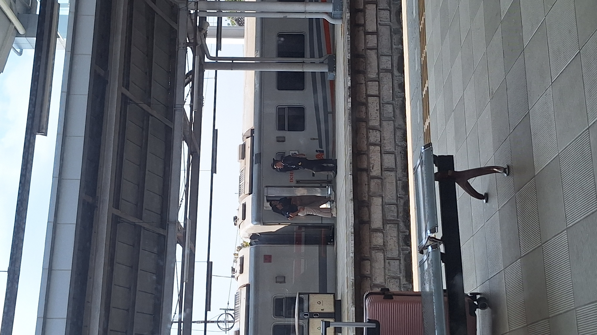 At a train station in Cikarang, Indonesia, a train stands adjacent to the platform. Two individuals stand near the train's entrance; one appears to be an official or conductor in uniform. Luggage rests on the platform close to a seating area against the wall. Overhead, a metal structure covers the platform.