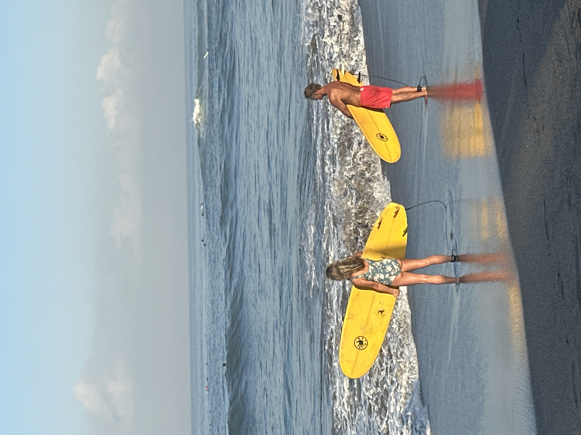 Two individuals, a woman and a man, stand on the wet sand at the edge of the ocean in North Kuta, Indonesia, each holding a yellow surfboard. The woman, facing away from the camera, wears a floral swimsuit and is positioned on the left side of the frame. The man, also facing away, is on the right, wearing red swim trunks. Both are ankle-deep in the shallow ocean water. The ocean stretches out to the horizon, with waves breaking closer to the shore. In the distance, several other surfers are visible in the water. The sky is a clear blue with a few wispy clouds. The sand in the foreground is dark and reflective, showing the shadows of the people and their surfboards.