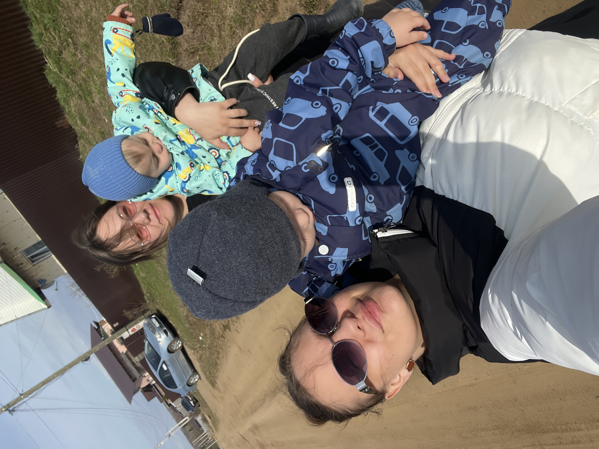The image shows a woman lying down outdoors with two young children. The woman is wearing sunglasses and a black and white jacket. One child, wearing a blue hat and a blue and green patterned jacket, is lying on her chest, and the other, wearing a grey hat and a blue car-patterned jacket, is also lying on her. They appear to be interacting closely, potentially cuddling.
In the background, there is a dirt road, a building with a brown roof, and a light blue car parked nearby. Overhead, there is a power line. The location is likely Ольховка, Russia.