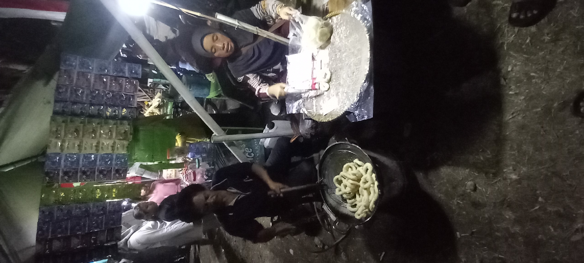 In Wanareja, Indonesia, a street vendor is frying snacks. A man, dressed in a black polo shirt and dark pants, is operating a large wok filled with golden-brown, ring-shaped snacks. The wok sits atop a makeshift stove. To his right, a woman wearing a dark hijab and a patterned shirt is preparing more of the snacks. She is sitting behind a table covered with aluminum foil and a woven mat, holding a plastic bag filled with raw dough. Behind the vendors, a stall displays rows of packaged goods. The scene takes place at night, illuminated by artificial lighting that casts shadows on the ground, which appears to be dirt and grass. Several other individuals are visible in the background, suggesting a bustling market or street food area.