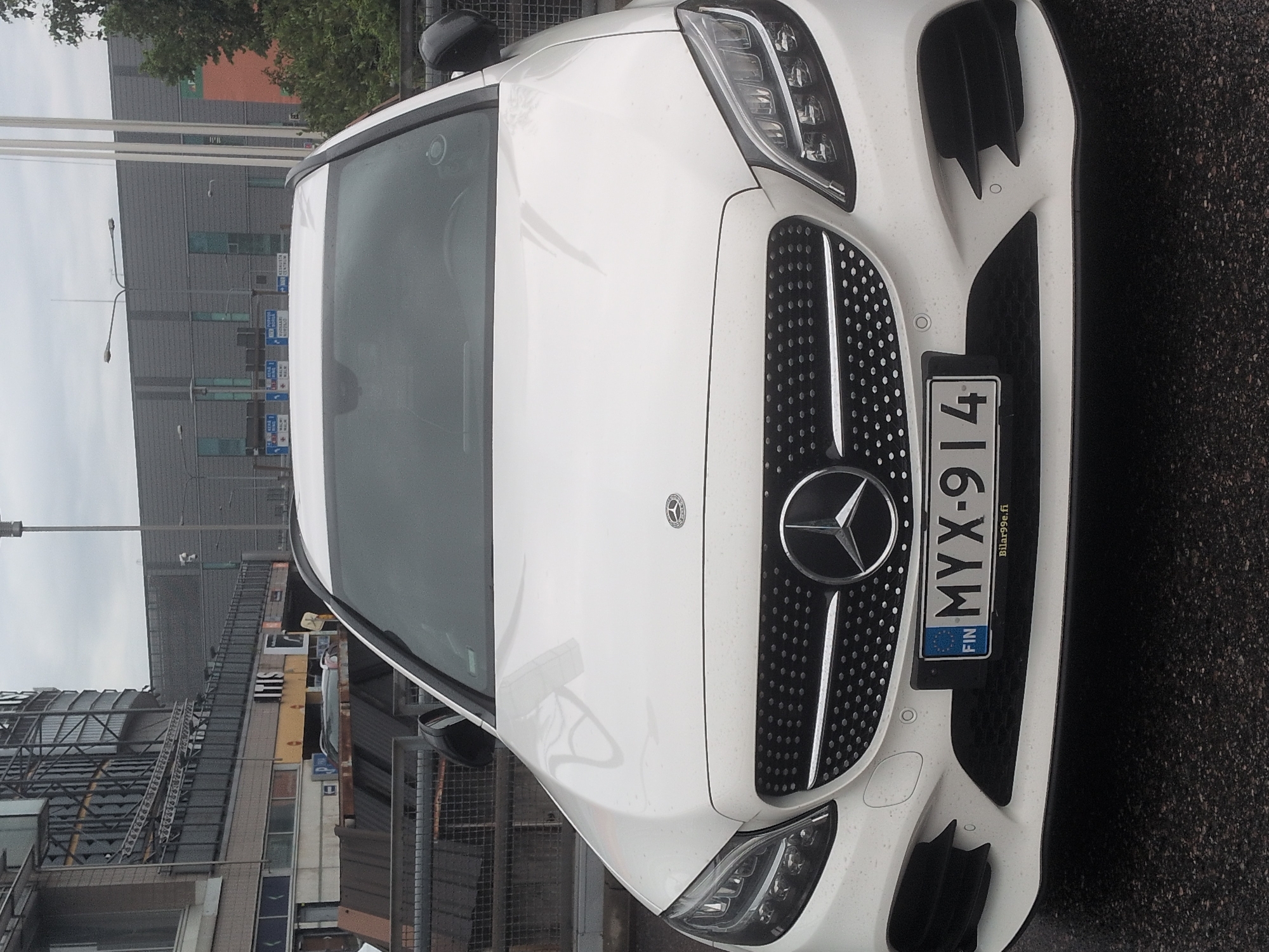 A white Mercedes-Benz C-Class sedan is parked on a wet, asphalt surface in front of a large, multi-story building with glass facades and metal scaffolding. The car's license plate reads "MYX-914" and has "FIN" on the left, indicating it is registered in Finland. A website address "Bilar99e.fi" is visible below the license plate. The building appears to be a commercial or public space, with signage for "ITIS" visible on a section of the building. The sky is overcast, suggesting a cloudy day. The image focuses on the front of the car, with the background showing urban infrastructure.